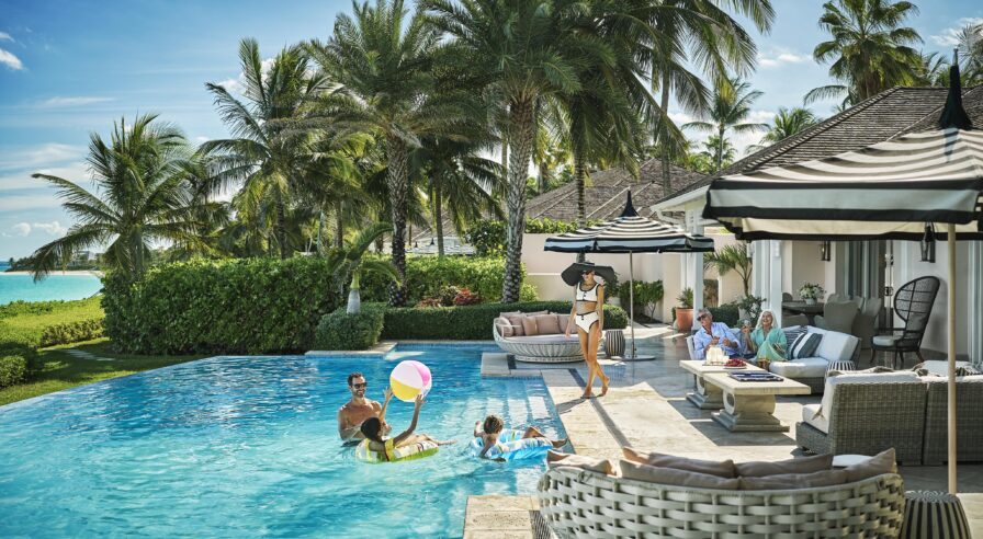 People enjoying a sunny day by an exclusive pool on their Ocean Club, A Four Seasons Resort and Residence villa. Some are swimming and playing with a beach ball, while others relax on the patio. Palm trees and a clear blue sky are in the background.