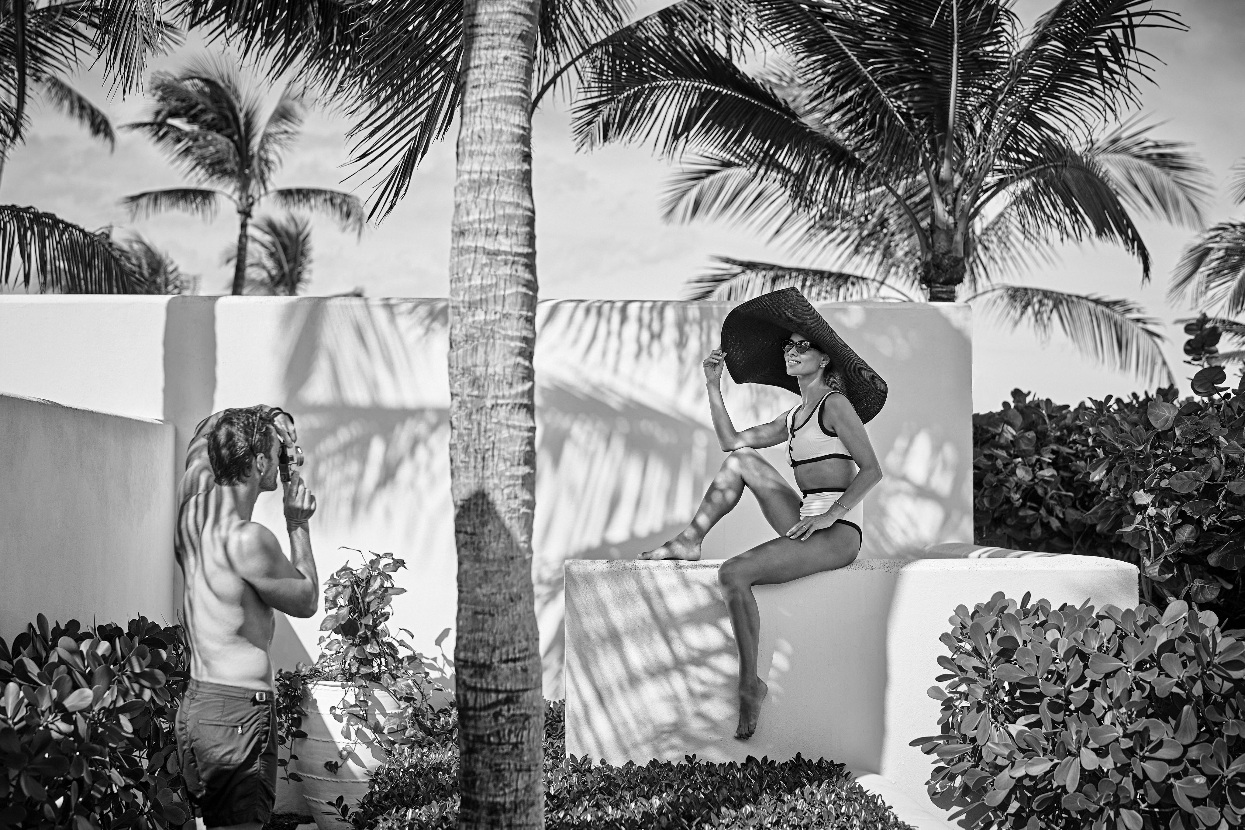 A woman in a large hat poses on a Ocean Club, A Four Seasons Resort property wall, while a man takes her photo. They are surrounded by tropical plants and palm trees in a bright outdoor setting.