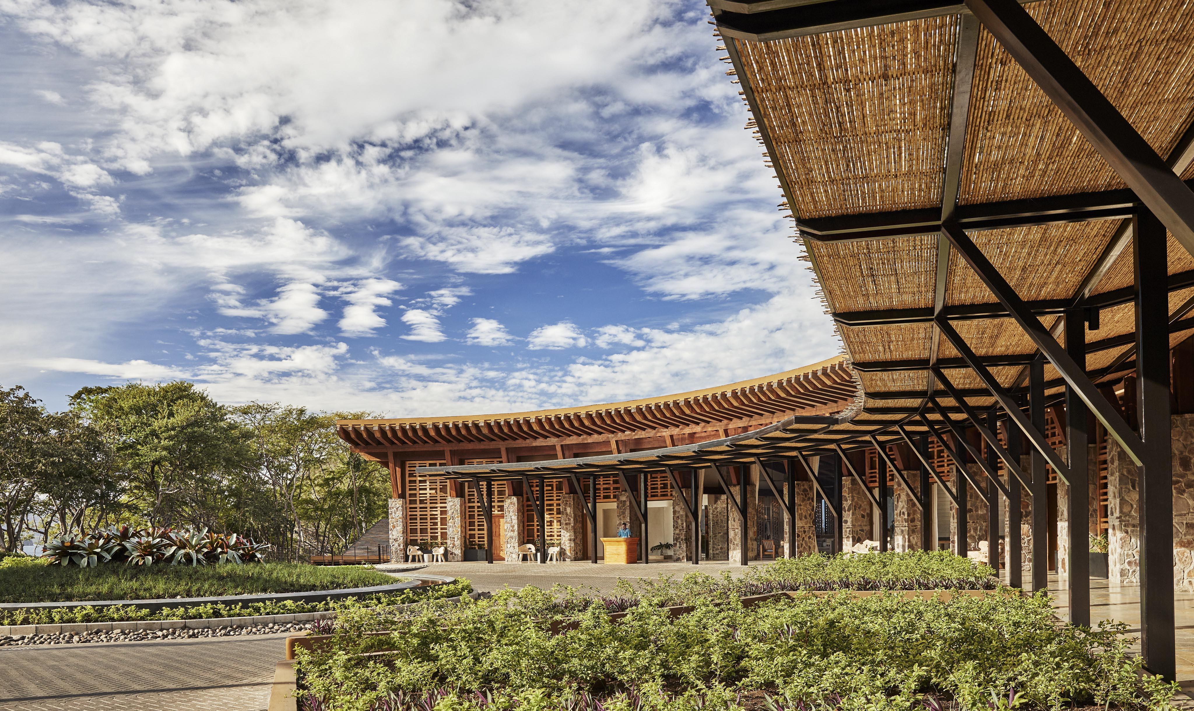 A wooden building with an open, curved design and woven roof panels evokes the architectural style of Central America, nestled among lush greenery and plants under a blue sky with scattered clouds.
