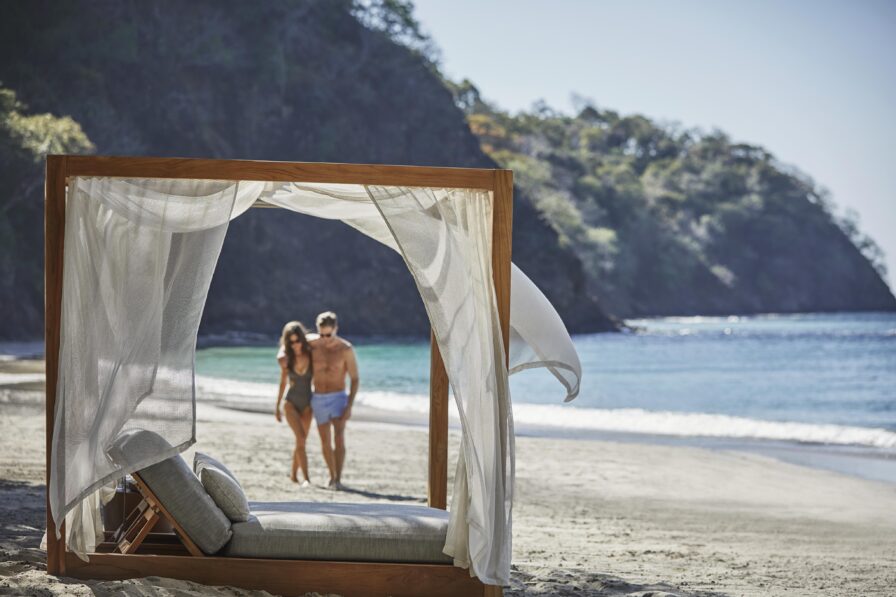 A couple strolls along a sandy beach near a wooden cabana with draped white fabric, capturing the essence of Central America's stunning landscapes. Hills and lush greenery rise in the background under a clear sky, offering a perfect blend of natural beauty and tranquility.