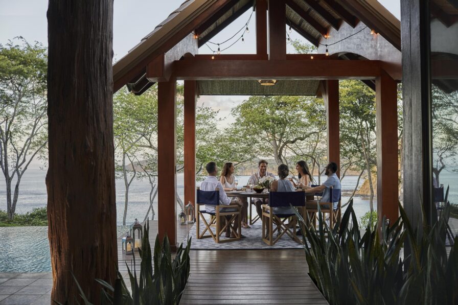 Five people are dining at a table in a pavilion overlooking a breathtaking outdoor view reminiscent of Central America's lush greenery and tranquil waters.