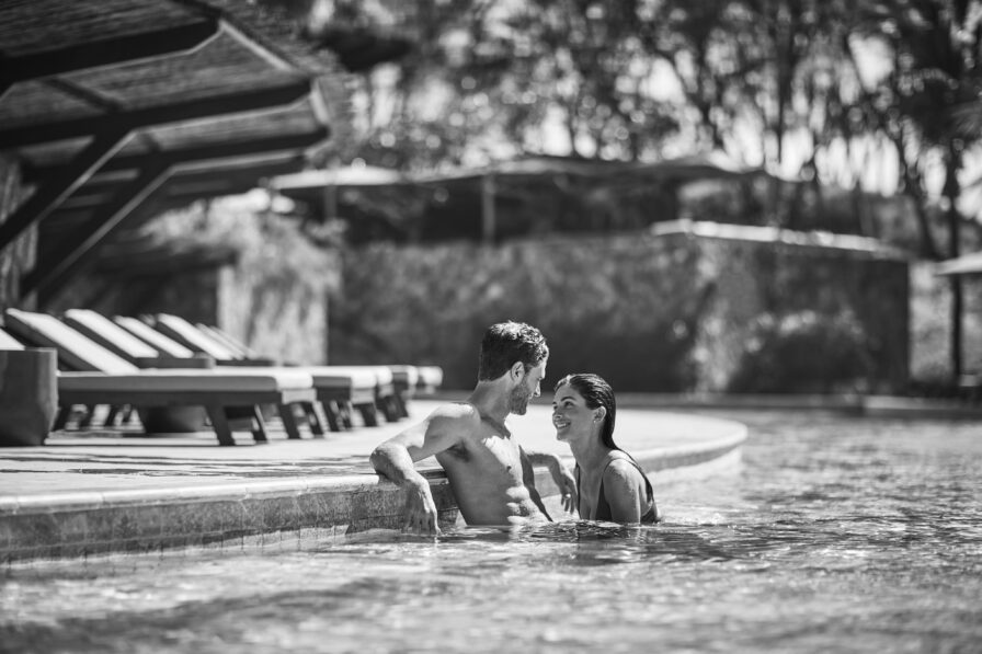 A man and woman smile at each other in a swimming pool, with deck chairs and tropical trees reminiscent of Central America in the background. Black and white image.