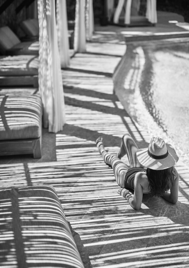 A person in a swimsuit lies by the pool in a tranquil scene reminiscent of South America's luxurious retreats, wearing a sun hat. Striped shadows from curtains create patterns on the ground and water, captured beautifully in this black and white image.