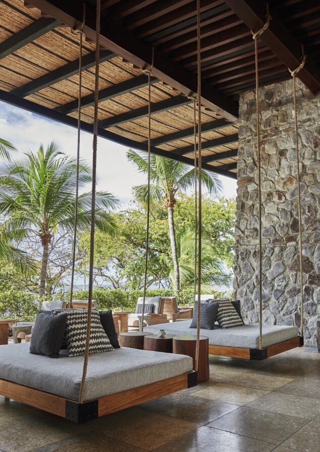 Outdoor patio with hanging beds, stone wall, and palm trees reminiscent of Central America. Cushions and pillows are placed on the beds, while a wooden ceiling provides shade.