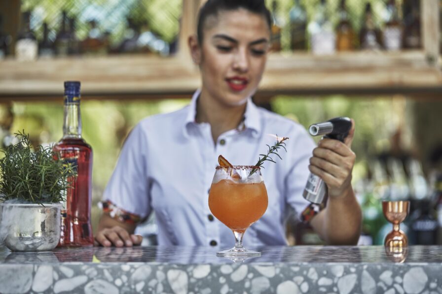 On a marble counter, a bartender from Central America expertly prepares a cocktail with a garnish, skillfully using a culinary torch. A bottle and plant add an elegant touch to her creative workspace.