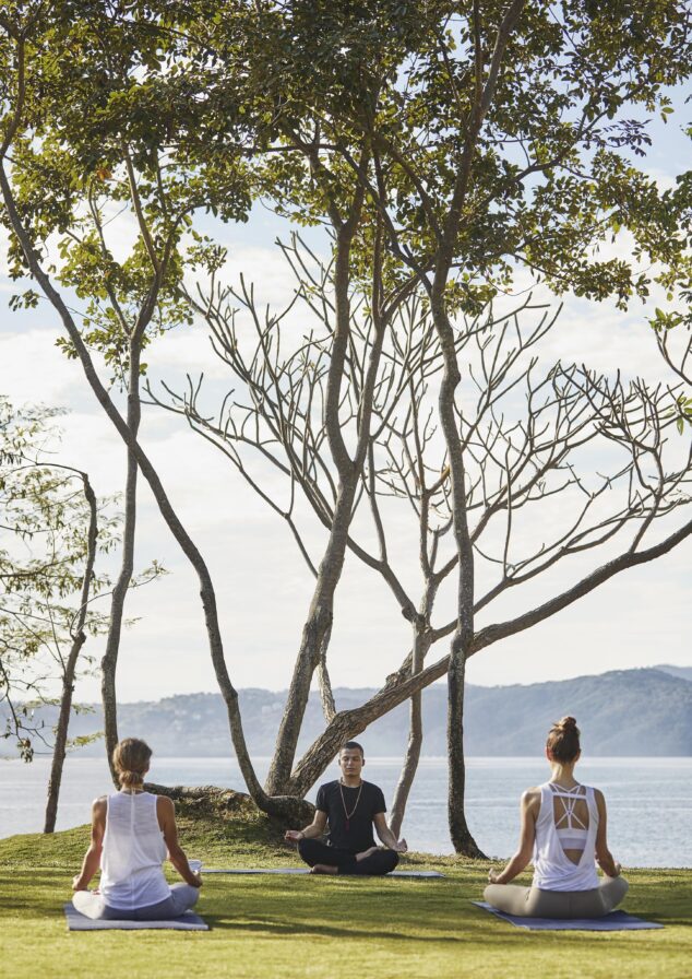 Three people are meditating on a grassy hill by the ocean, surrounded by lush trees typical of Central America, under a clear sky.