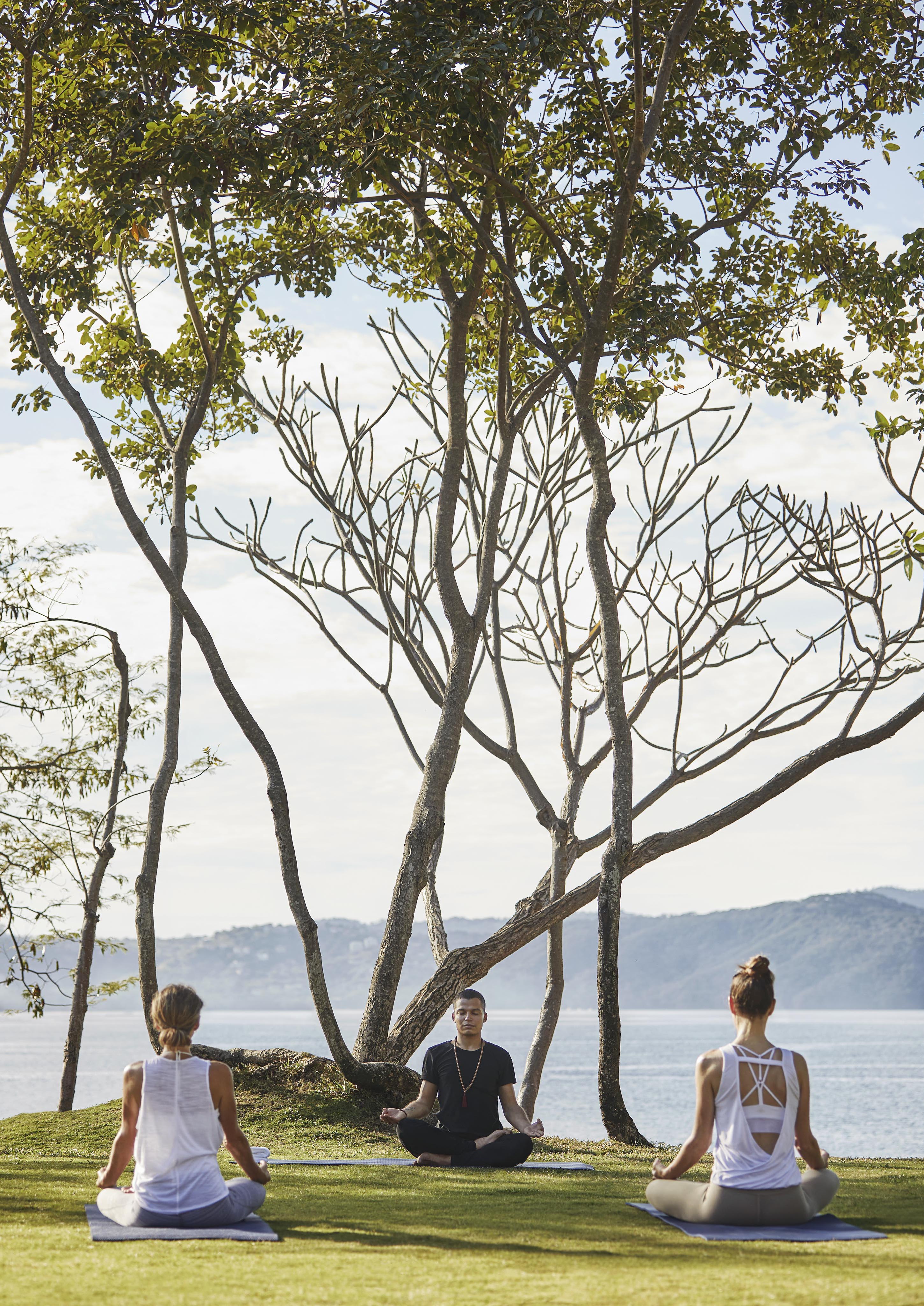 Three people are meditating on a grassy hill by the ocean, surrounded by lush trees typical of Central America, under a clear sky.