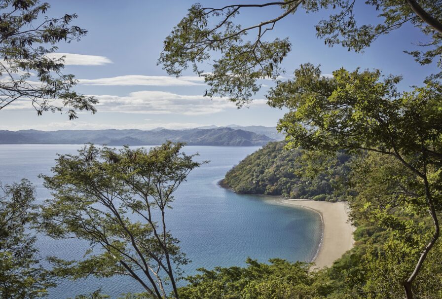 Aerial view of a secluded beach in South America, surrounded by lush greenery and calm blue water, with a backdrop of distant mountains under a clear sky.