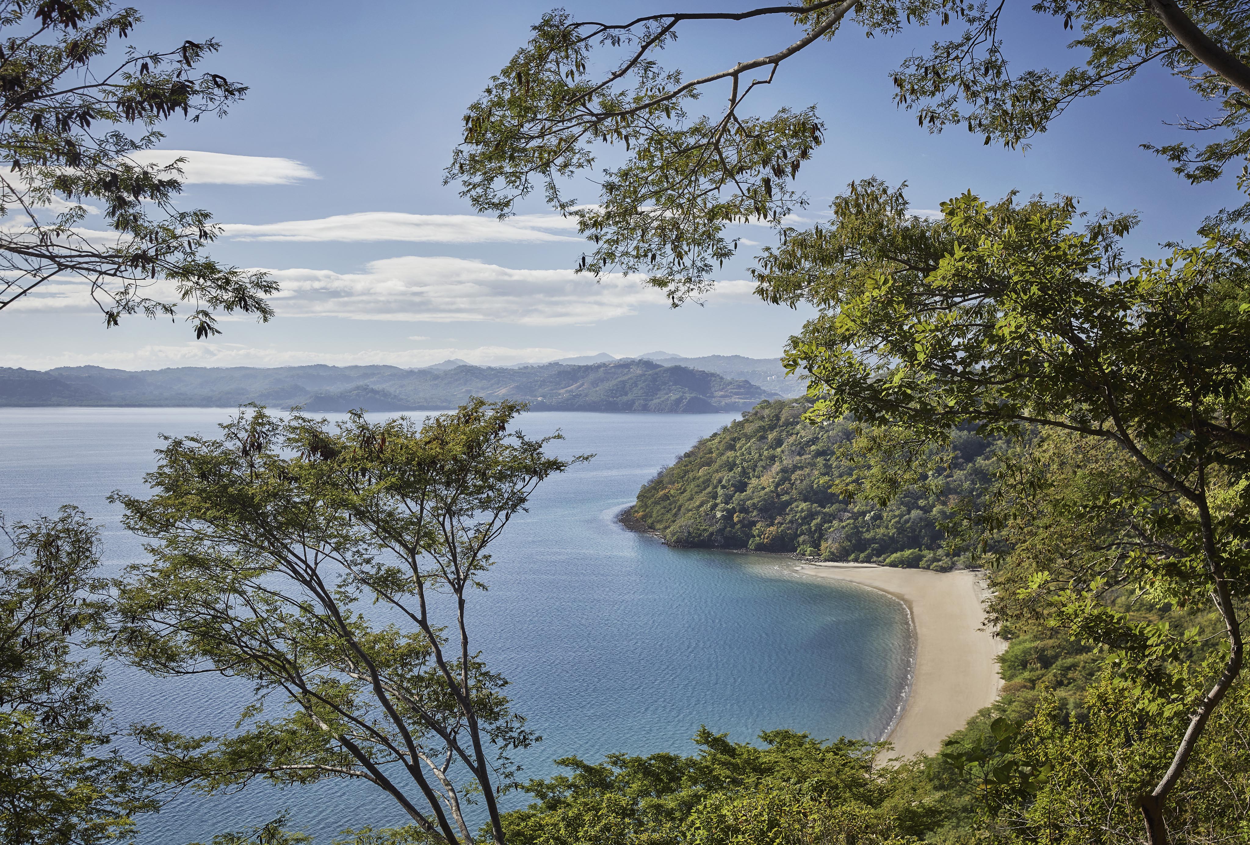 Aerial view of a secluded beach in South America, surrounded by lush greenery and calm blue water, with a backdrop of distant mountains under a clear sky.