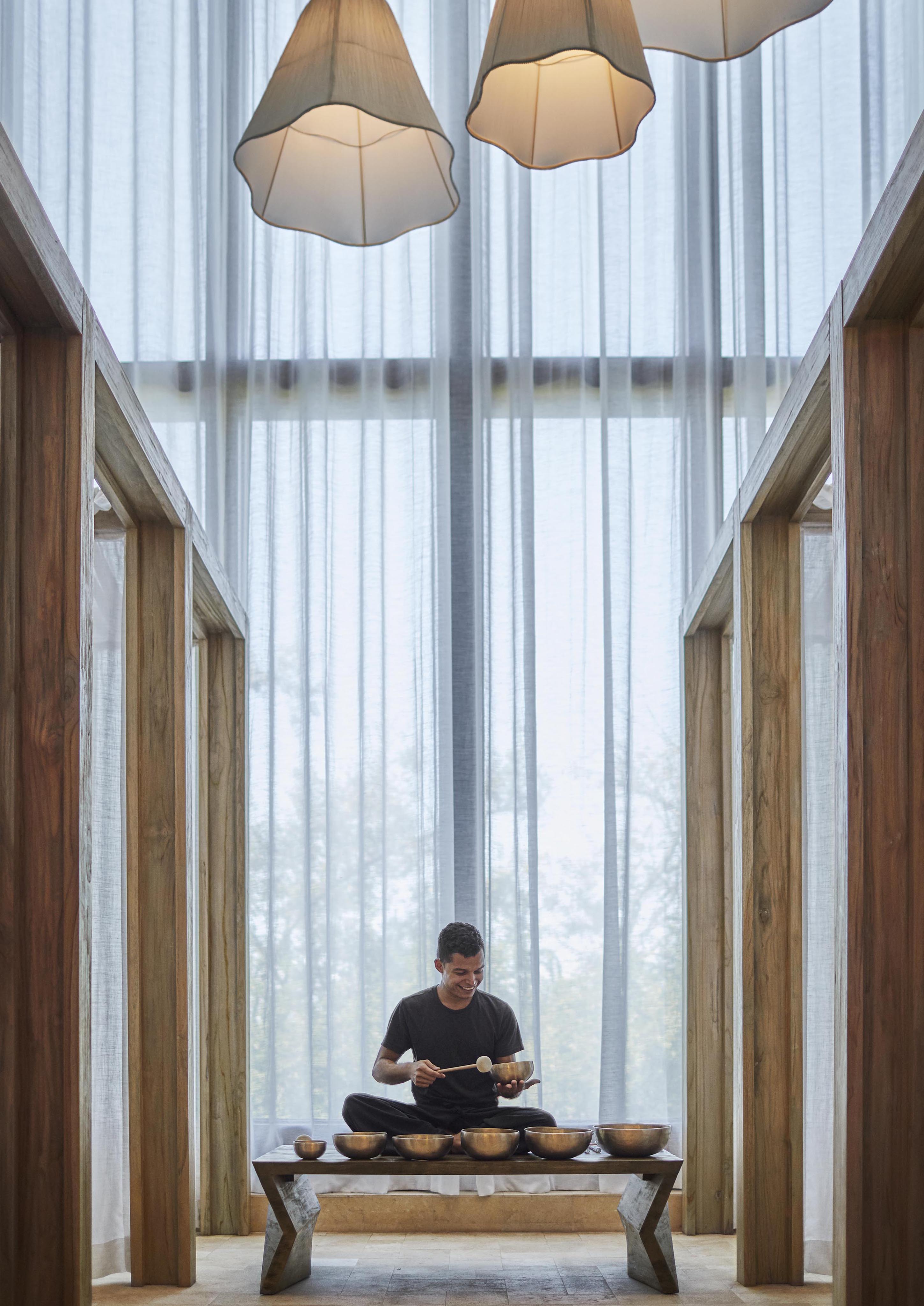 A person sits on a wooden bench, playing singing bowls with mallets in a well-lit room adorned with wooden structures and sheer curtains, evoking the serene ambiance of Central America's natural beauty.