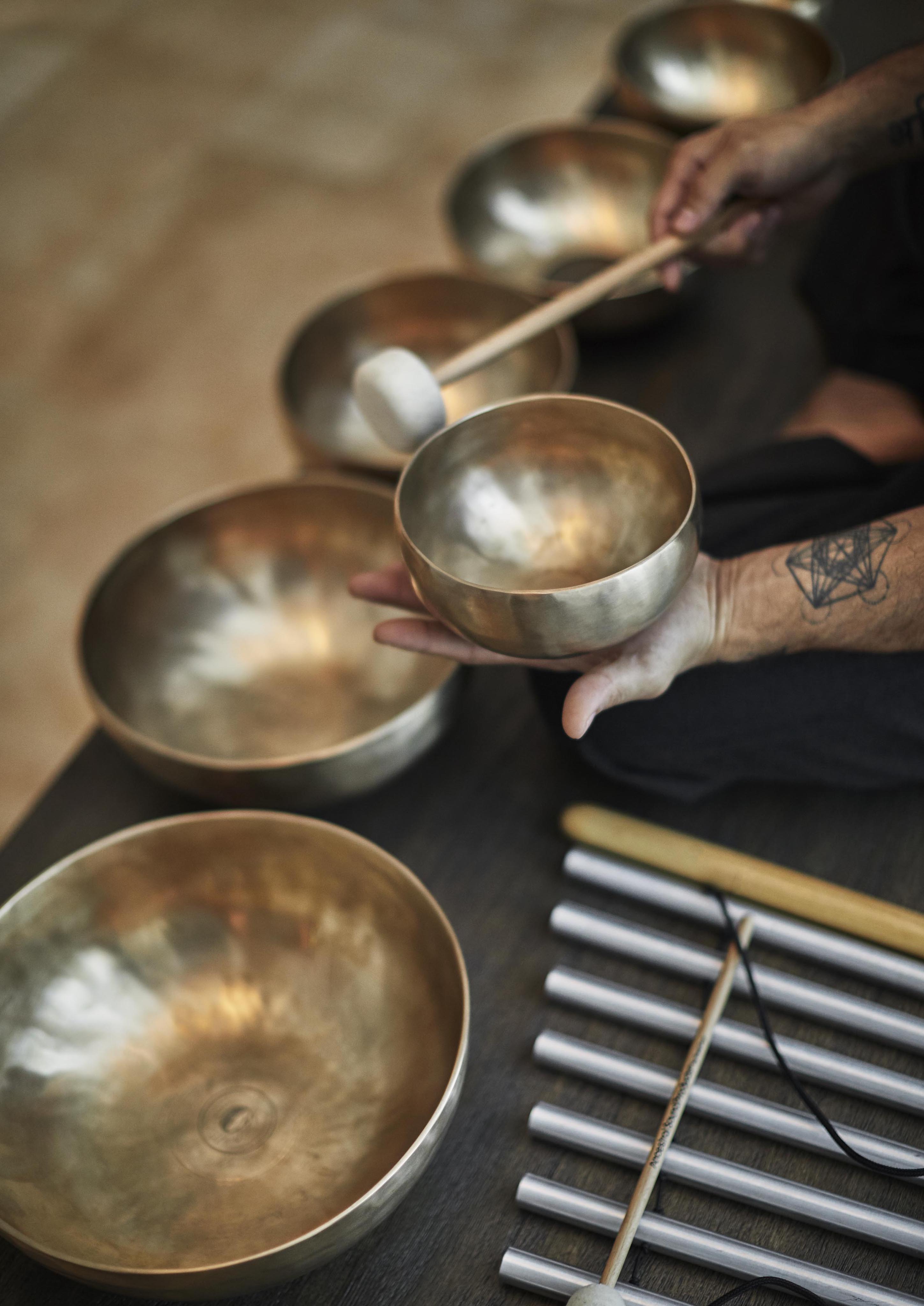 A person holding a mallet taps a large singing bowl, surrounded by other bowls and tuning forks, suggesting a sound healing session reminiscent of ancient practices in Central America.
