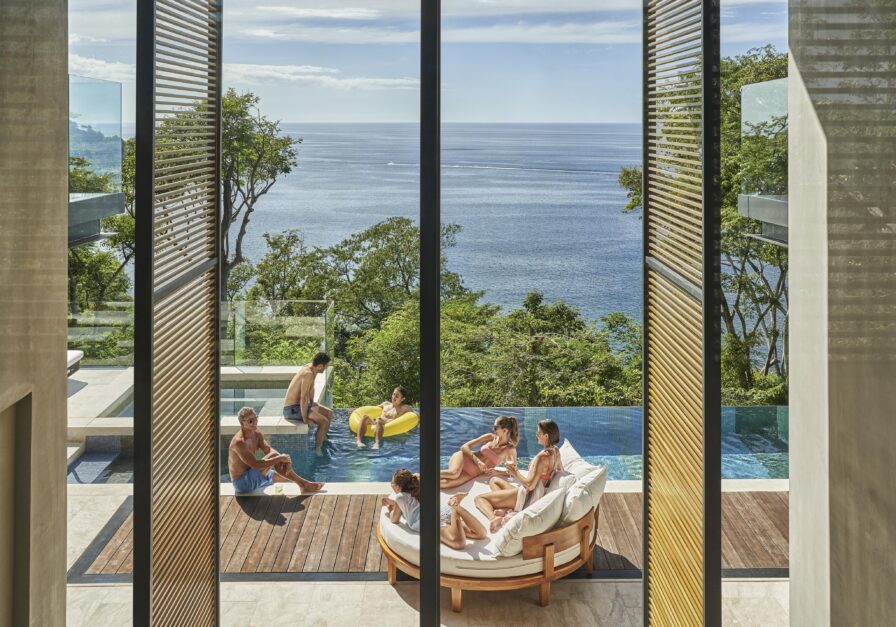 People relax in and around a pool overlooking the ocean, evoking the vibrant spirit of Central America. Some sit on lounge chairs, while others use a yellow float. Trees and clear skies are visible in the background, enhancing this idyllic scene.