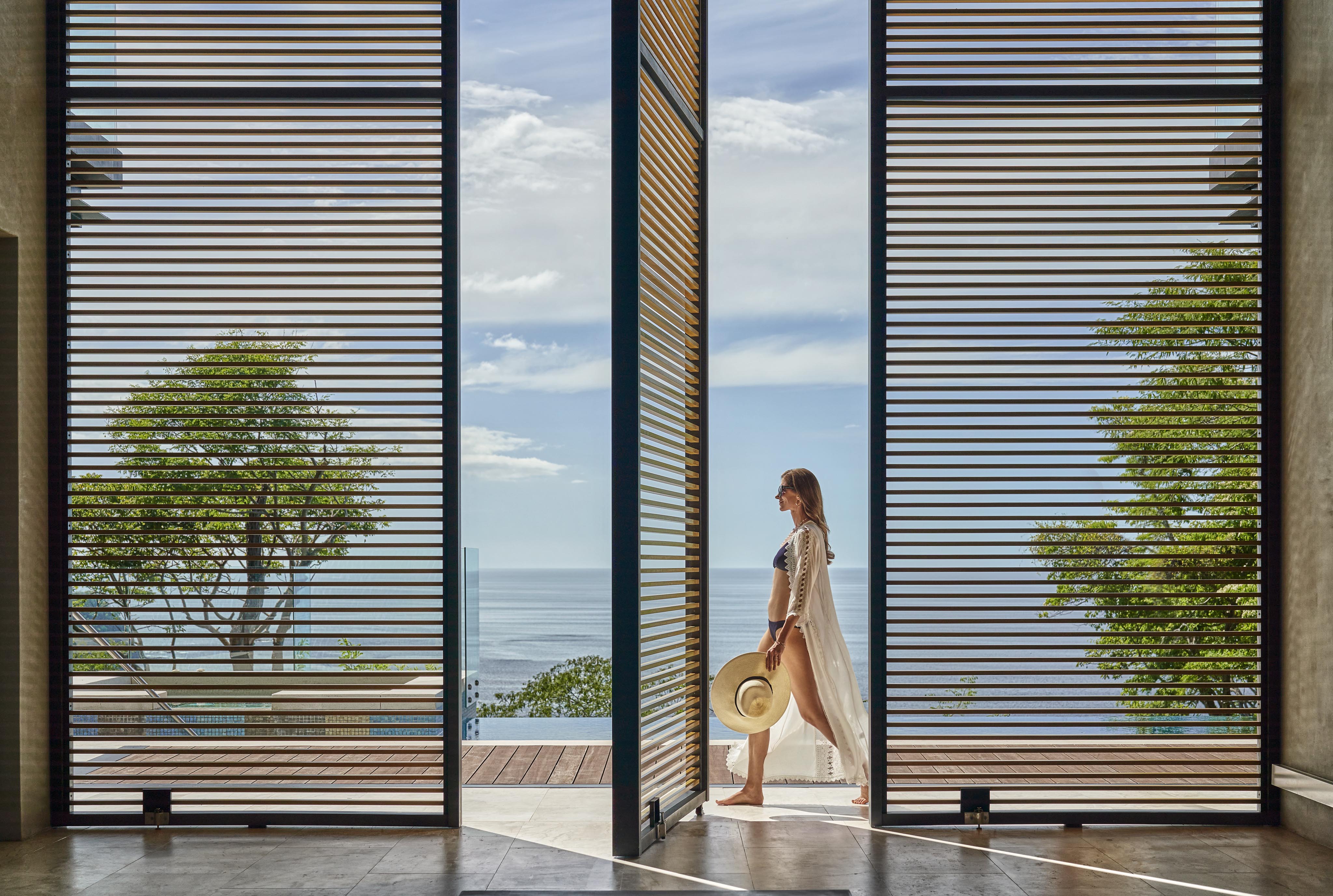 A woman holding a hat strolls along a wooden deck through large open sliding doors with a slatted design, embracing the tranquil ambiance of Central America. Ocean and lush trees paint the serene backdrop, capturing a perfect blend of nature and architecture.
