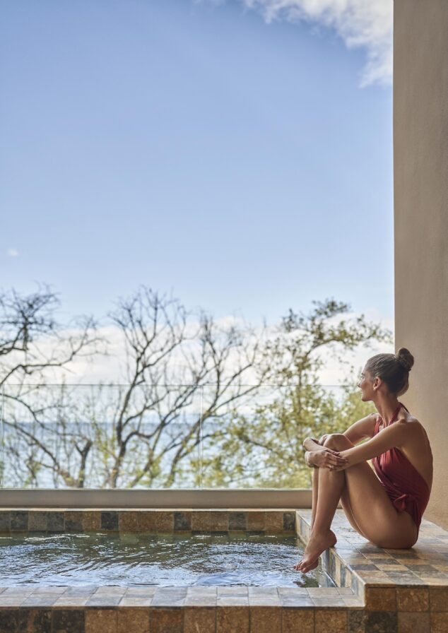Woman in a red swimsuit sitting on the edge of an outdoor hot tub, gazing at the scenic views reminiscent of Central America, with lush trees and a cloudy sky enhancing the tranquil setting.