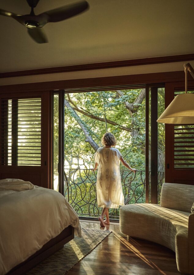 A person in a light robe stands on a balcony, gazing at the lush trees of Central America. A bed and chair rest in the foreground, with sliding doors open to the vibrant outside greenery.