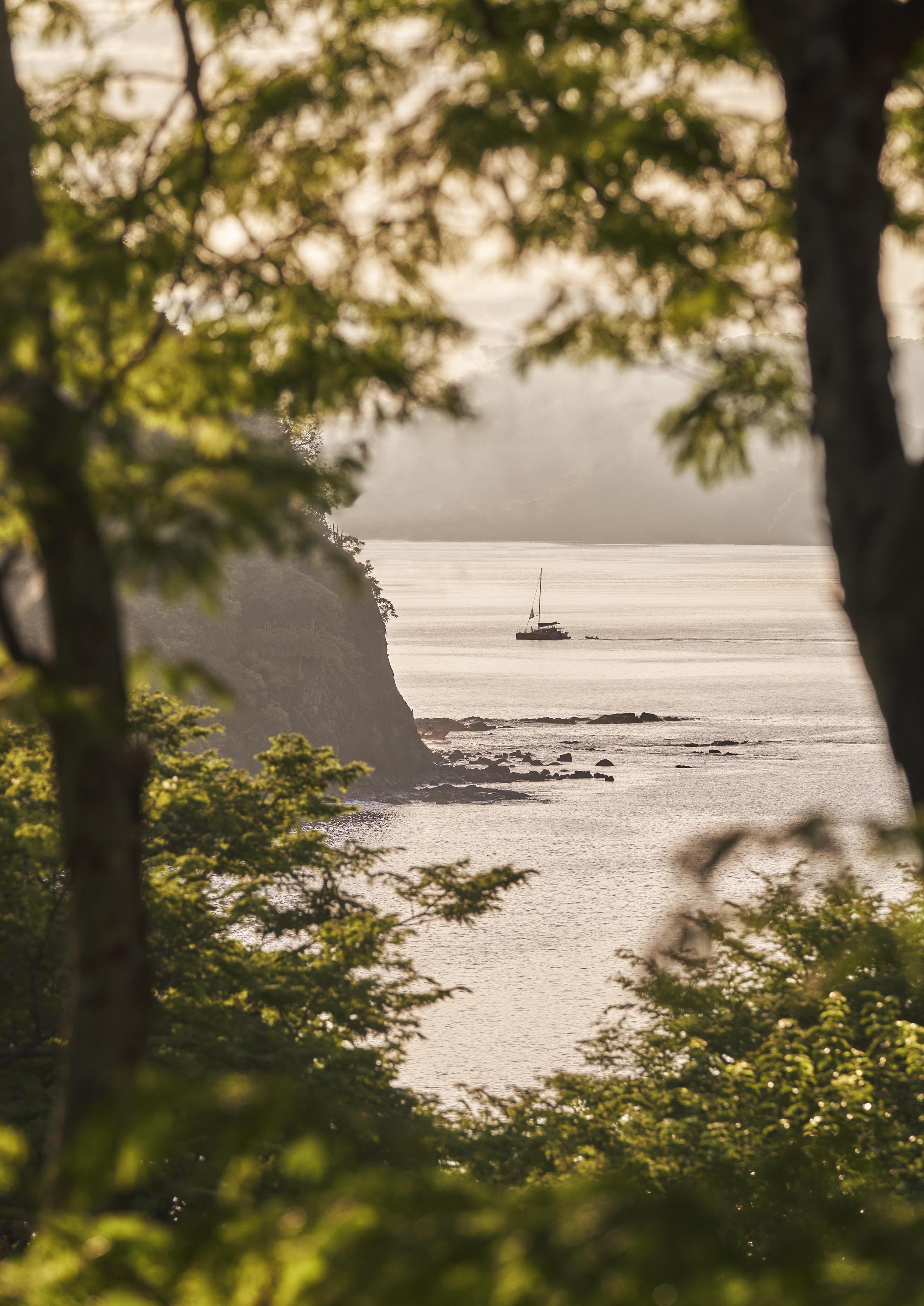 A sailboat is anchored near a rocky Central American coast, framed by lush green trees in the foreground, with sunlight casting a warm glow over the water and landscape.