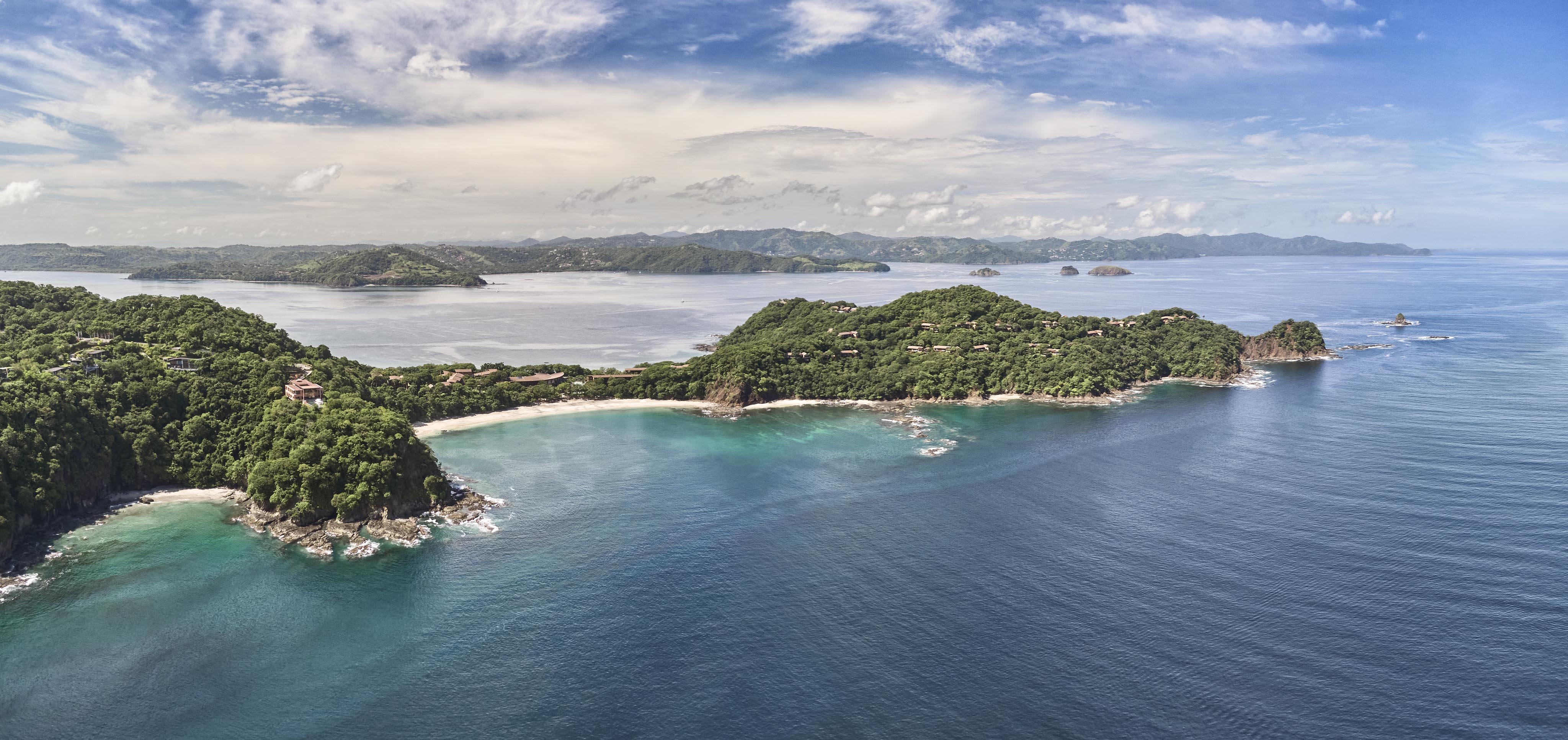 Aerial view of a coastal landscape in Central America, featuring lush green hills, sandy beaches, and clear blue ocean under a partly cloudy sky.