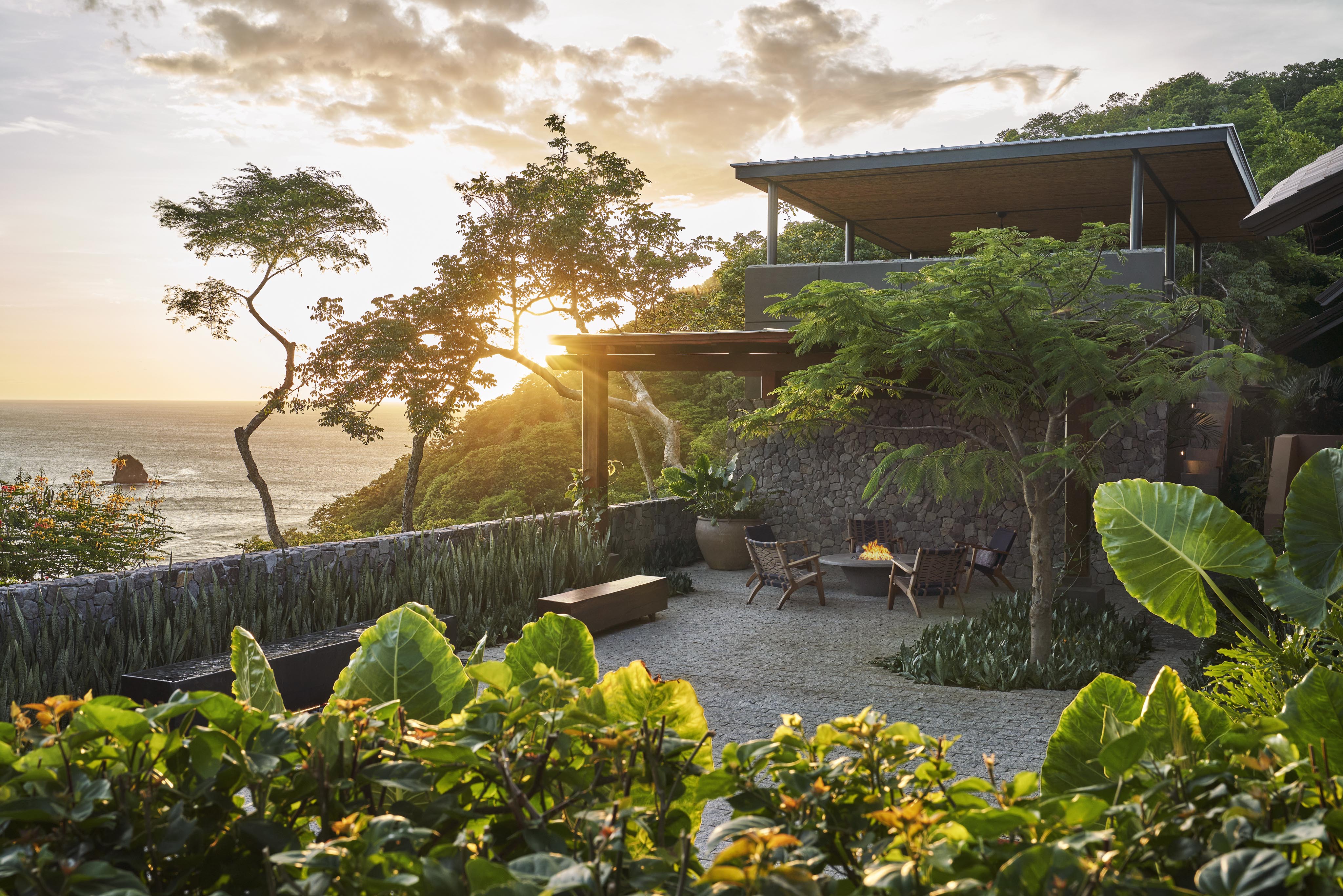 A modern house with a stone exterior overlooks the ocean at sunset, nestled amidst lush greenery and trees, reflective of the vibrant landscapes of Central America. A cozy seating area in the foreground invites relaxation.