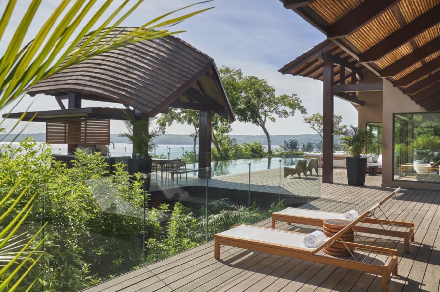 A luxurious outdoor deck featuring two wooden lounge chairs with white towels, surrounded by lush greenery, overlooking a pool and a scenic ocean view under the clear skies of Central America.