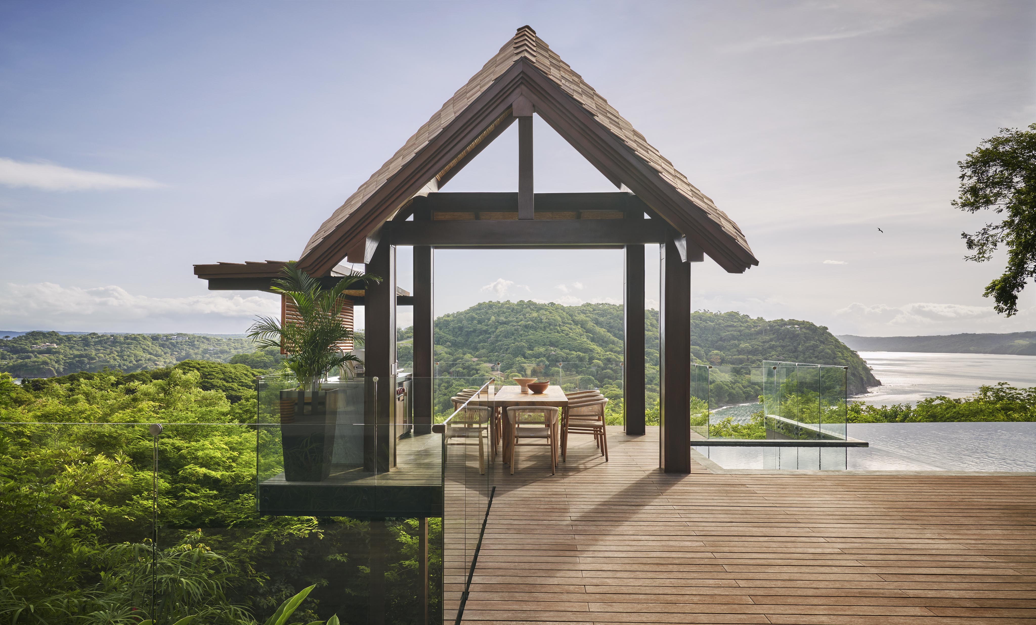 A wooden deck with a table and chairs sits under a peaked roof, offering panoramic views reminiscent of Central America's lush green hills and shimmering waters. A glass railing surrounds the deck, ensuring an unobstructed scenic vista.