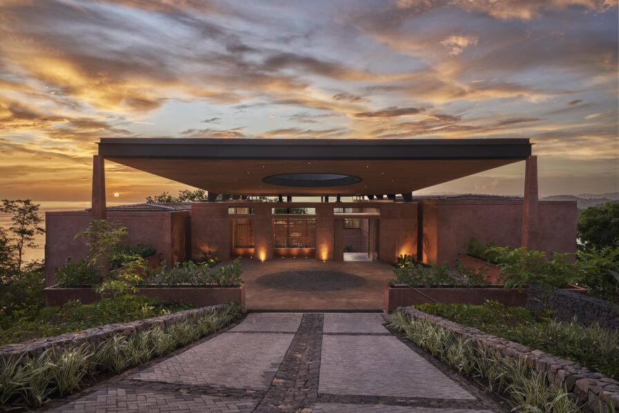 A modern house with a flat roof and central circular opening is nestled amidst lush plants, offering stunning ocean views at sunset—a scene reminiscent of tropical paradises in South America.