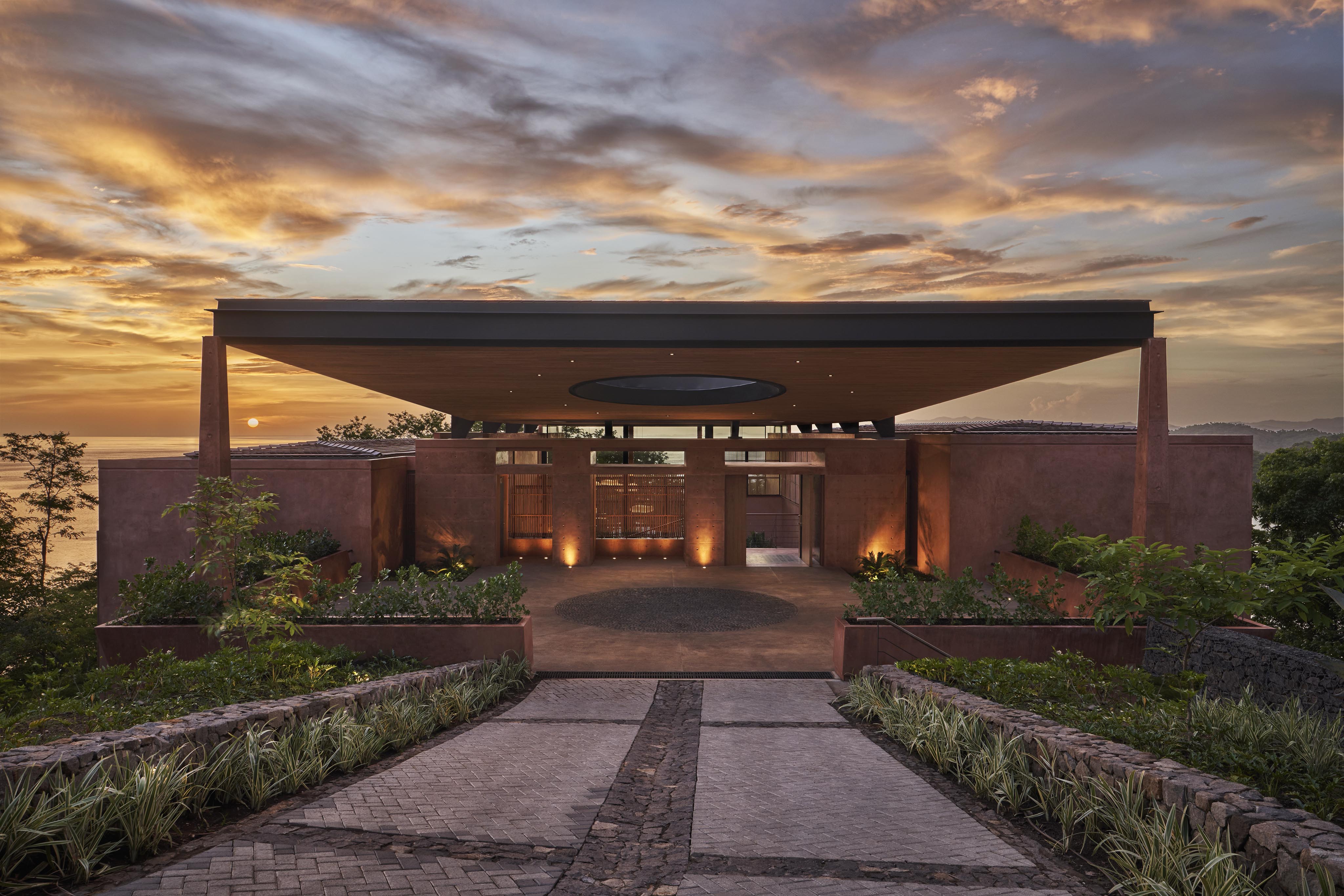 A modern house with a flat roof and central circular opening is nestled amidst lush plants, offering stunning ocean views at sunset—a scene reminiscent of tropical paradises in South America.