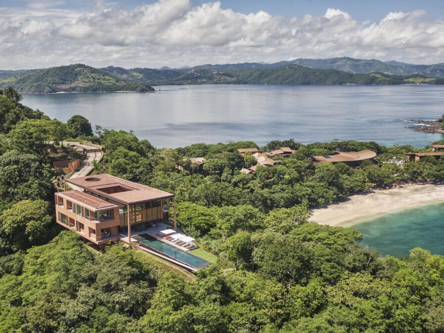 Modern house with a pool overlooking a lush green landscape and beach, nestled in the hilly terrain of Central America, under a partly cloudy sky.