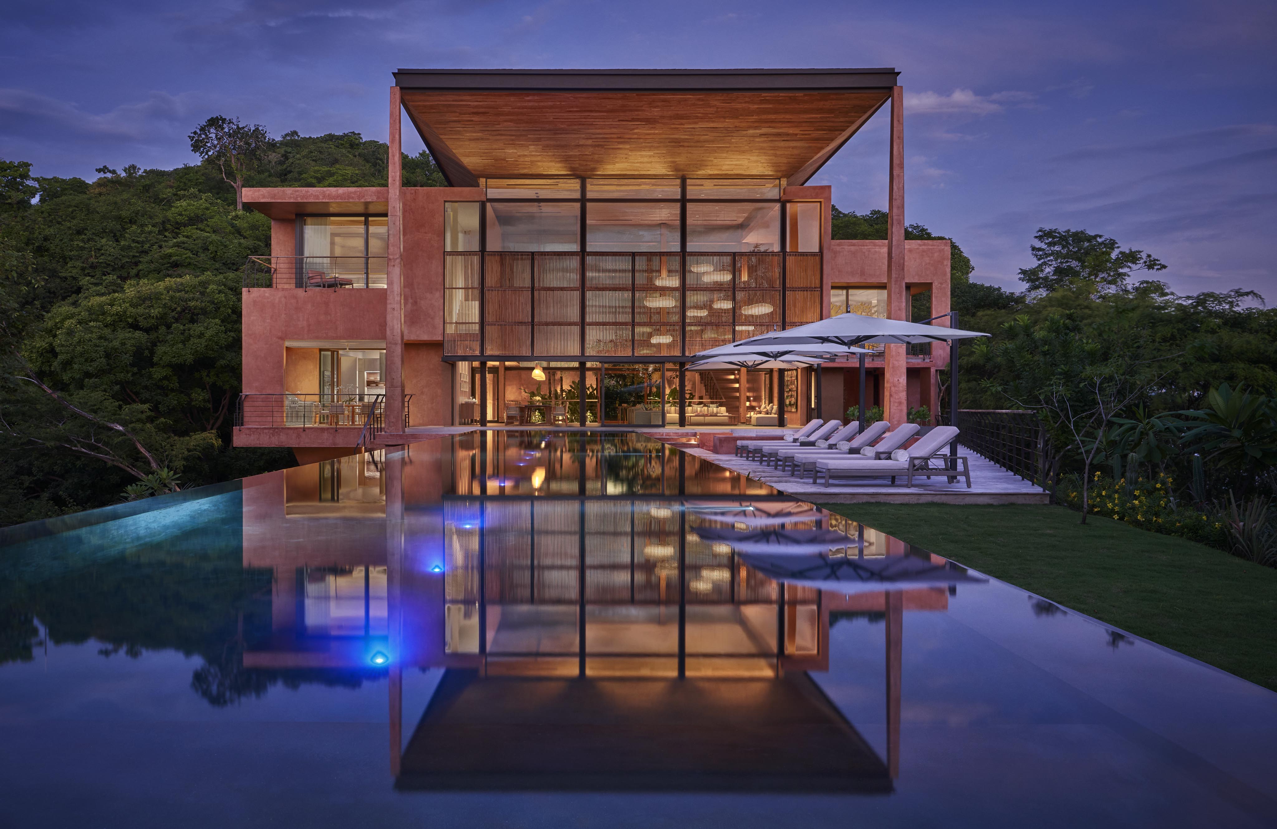 Modern, multi-story house with large glass windows and a pool reflecting the building. Surrounded by lush greenery reminiscent of Central America under an early evening sky.