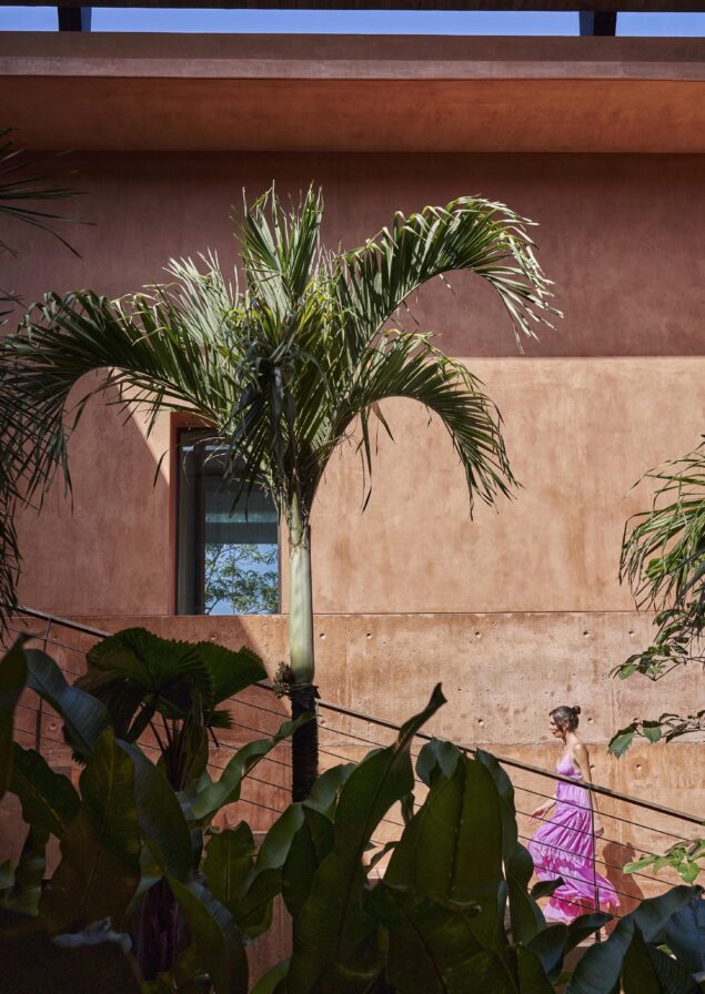 A person in a pink dress strolls along a railing against a terracotta wall, surrounded by lush plants and a palm tree, evoking the vibrant spirit of South America.