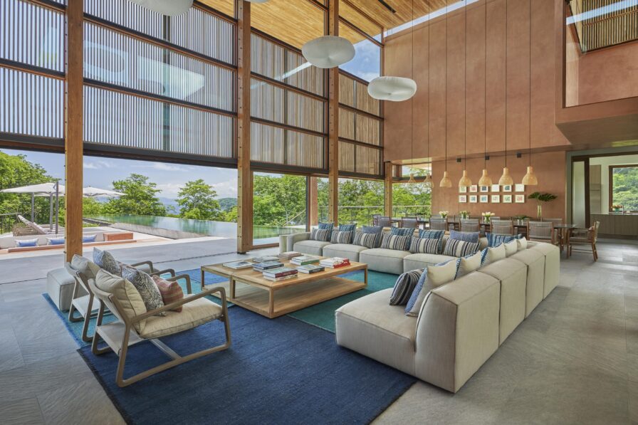 Spacious modern living room with large windows offers a touch of Central America's warmth, featuring a beige sectional sofa, armchairs, coffee table with books, pendant lights, dining table, and stunning poolside view.