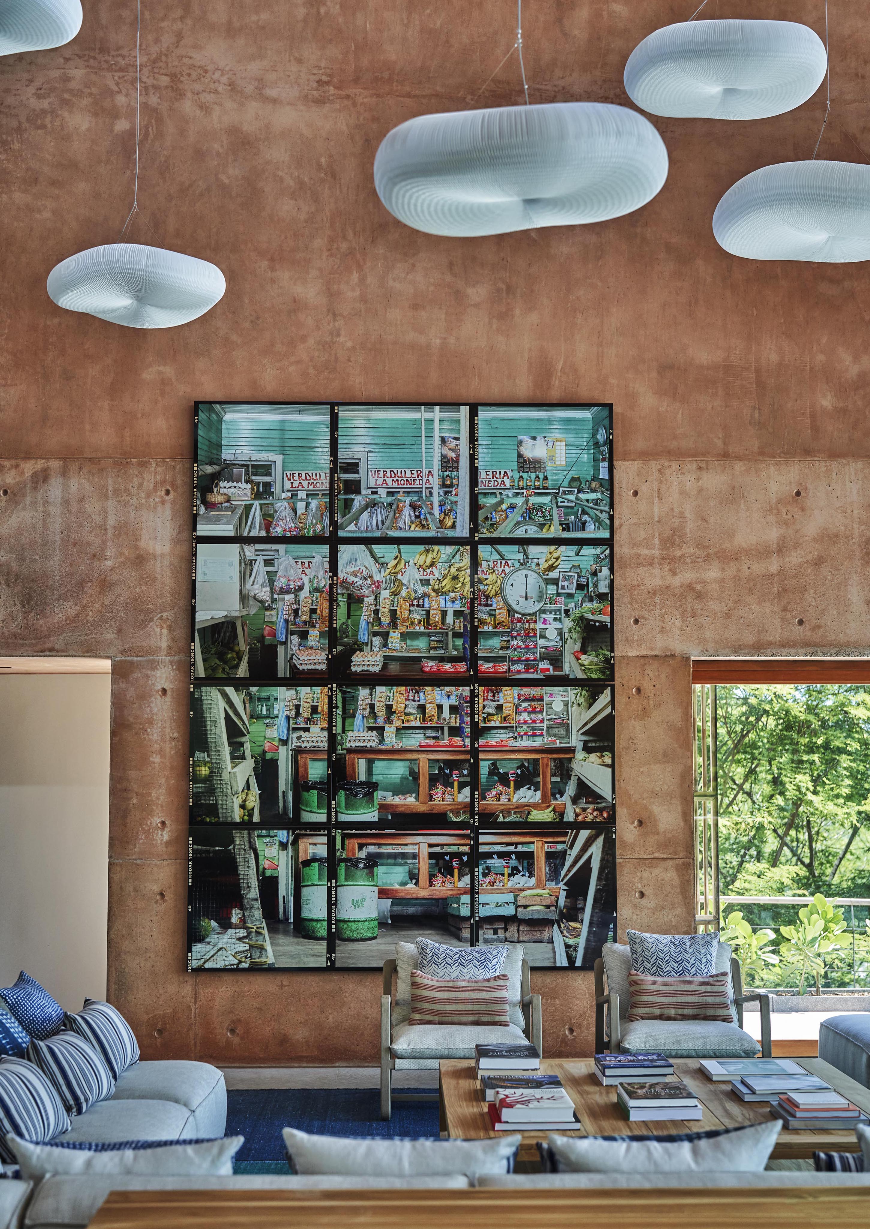 A modern living room features a large, colorful collage artwork inspired by South America on a textured wall. White, cloud-like light fixtures hang above a sofa and tables below. Large windows offer a stunning view outside.