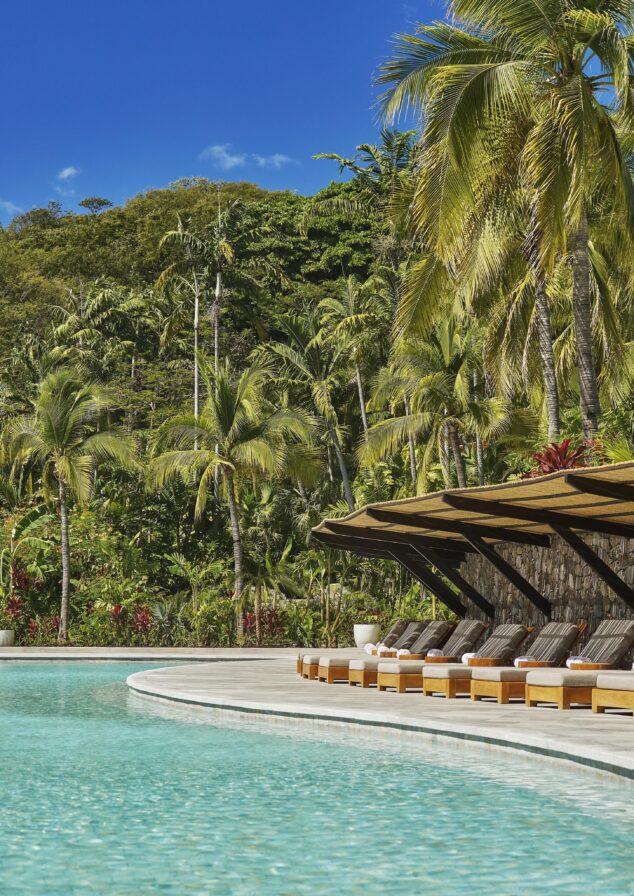 A serene outdoor swimming pool, reminiscent of a hidden oasis in Central America, is surrounded by palm trees and lush greenery under a clear blue sky. Sun loungers are perfectly lined up on the poolside, inviting relaxation amidst the tranquil beauty.