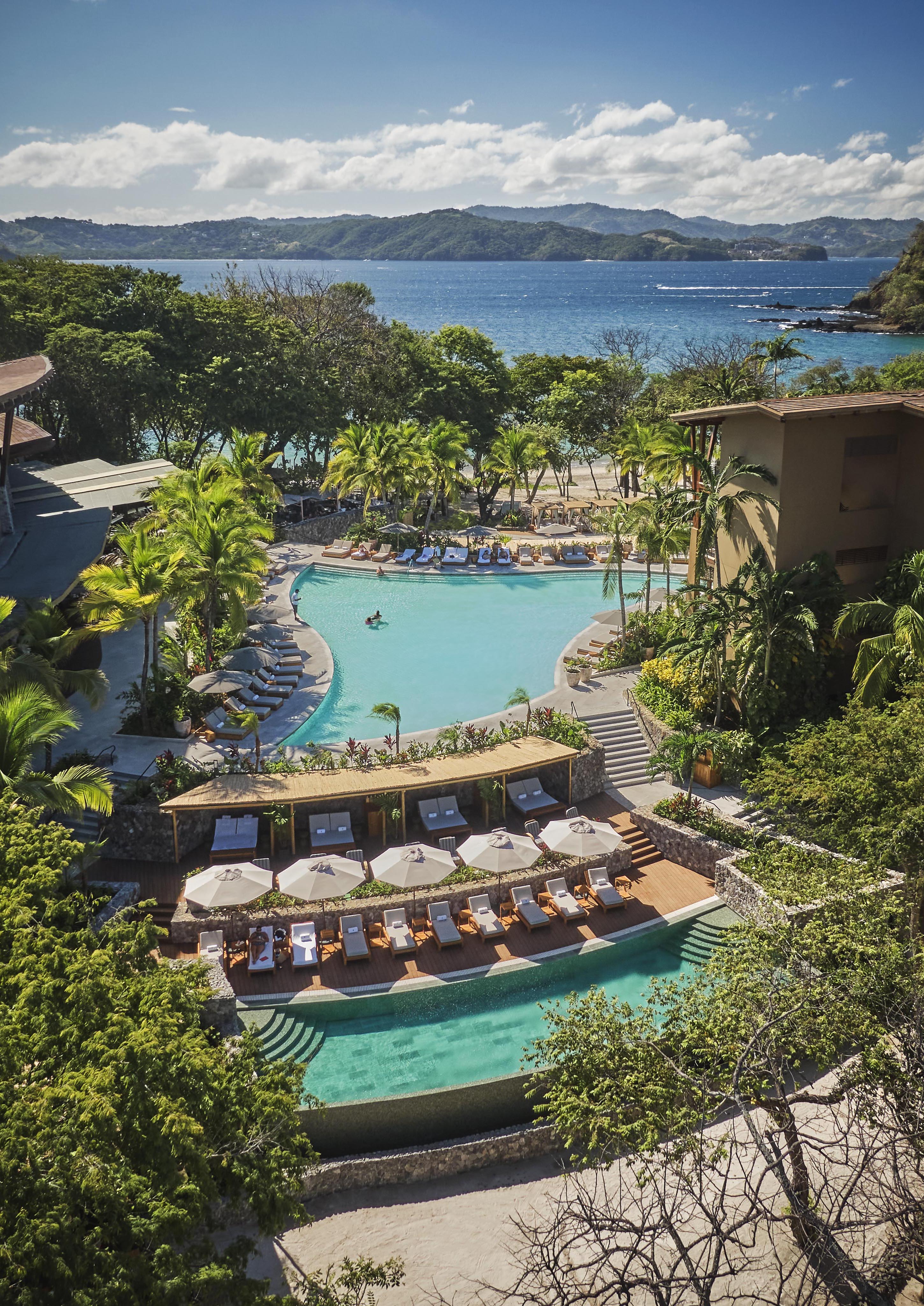 Aerial view of a Central American resort featuring a large pool surrounded by lounge chairs, palm trees, and buildings. The ocean is visible in the background under a partly cloudy sky.