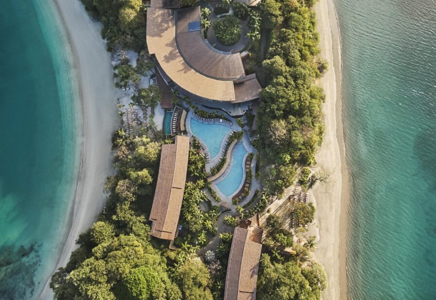Aerial view of a resort in Central America with several buildings and a pool surrounded by lush greenery, nestled between two sandy beaches and turquoise water.