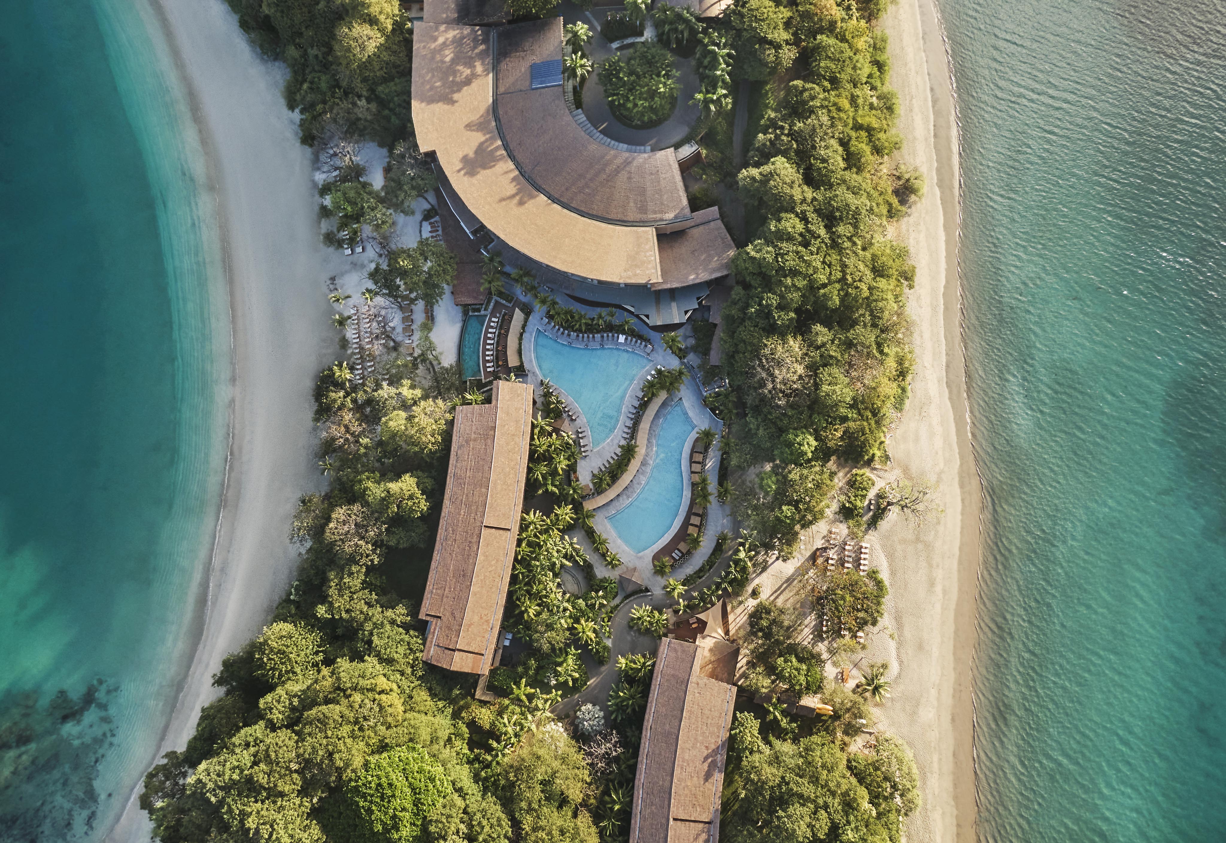 Aerial view of a resort in Central America with several buildings and a pool surrounded by lush greenery, nestled between two sandy beaches and turquoise water.
