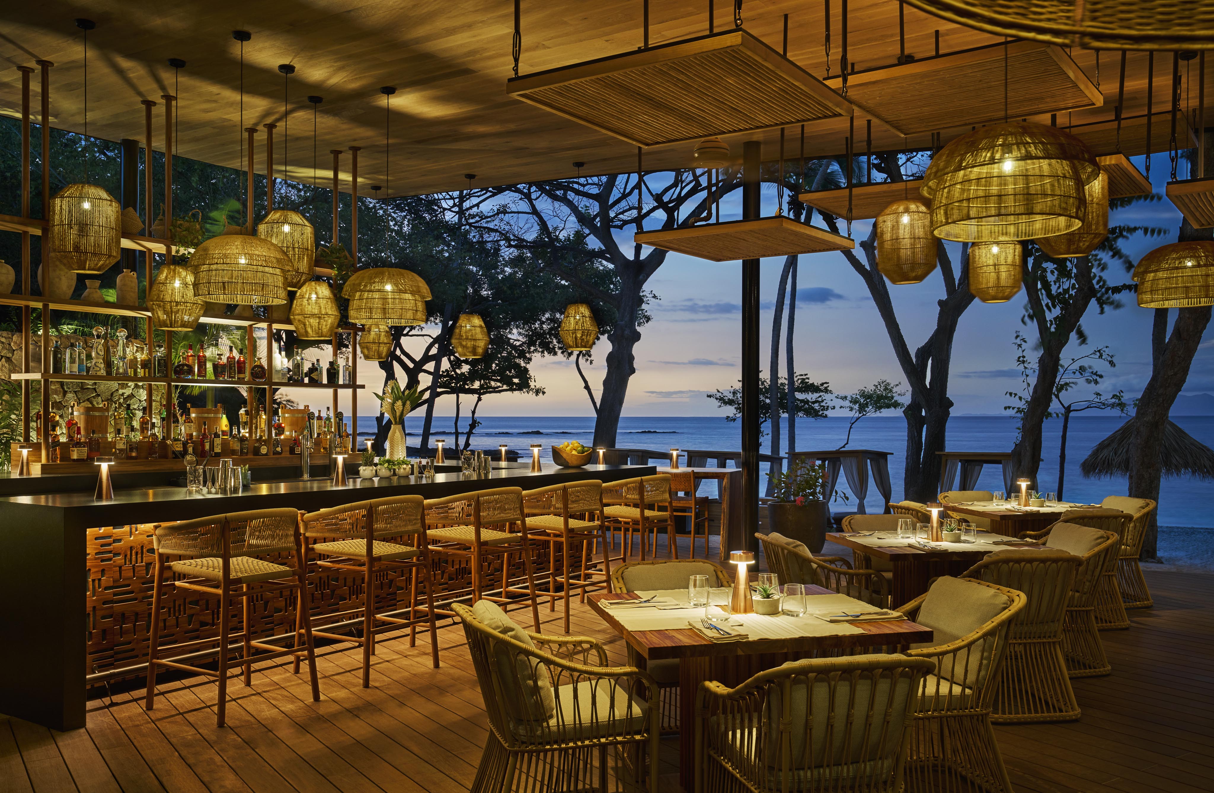 Outdoor bar and dining area at sunset, featuring wicker furniture, hanging woven light fixtures, and a view of the sea reminiscent of a vibrant Central American retreat.