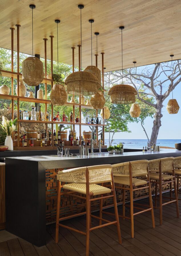 Discover a modern outdoor bar reminiscent of Central America's charm, featuring wicker lighting, wooden stools, and a stocked shelf set against a backdrop of trees and ocean.