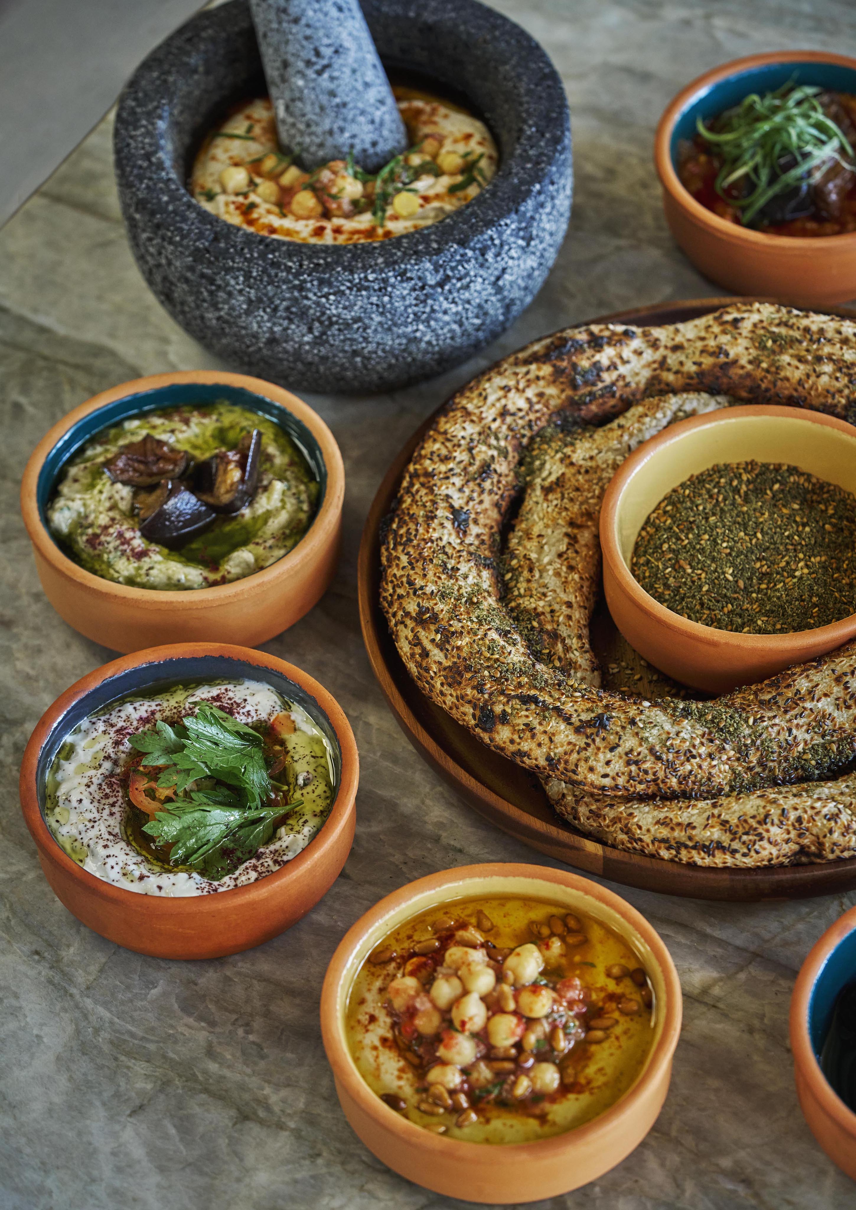 A variety of Middle Eastern dips in terracotta bowls, a stone mortar, and a large seeded bread ring grace the table with an unexpected flair reminiscent of South America's vibrant culinary traditions.