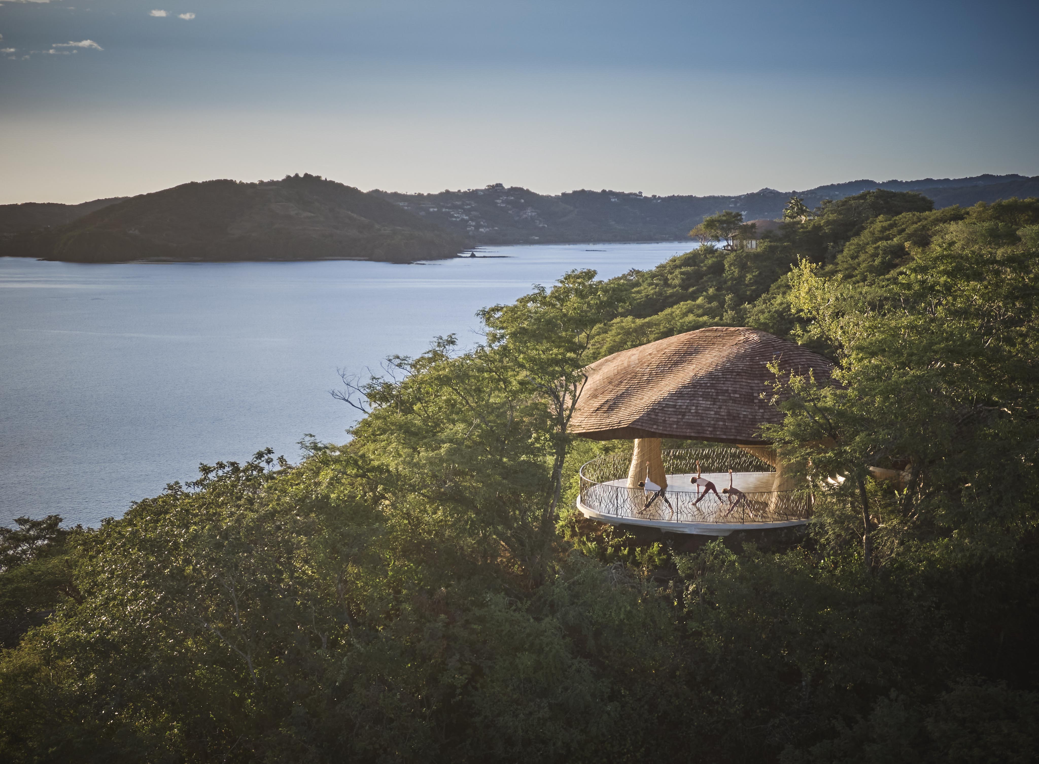 A thatched-roof pavilion nestled in lush greenery overlooks a calm body of water with the hills of Central America in the background.