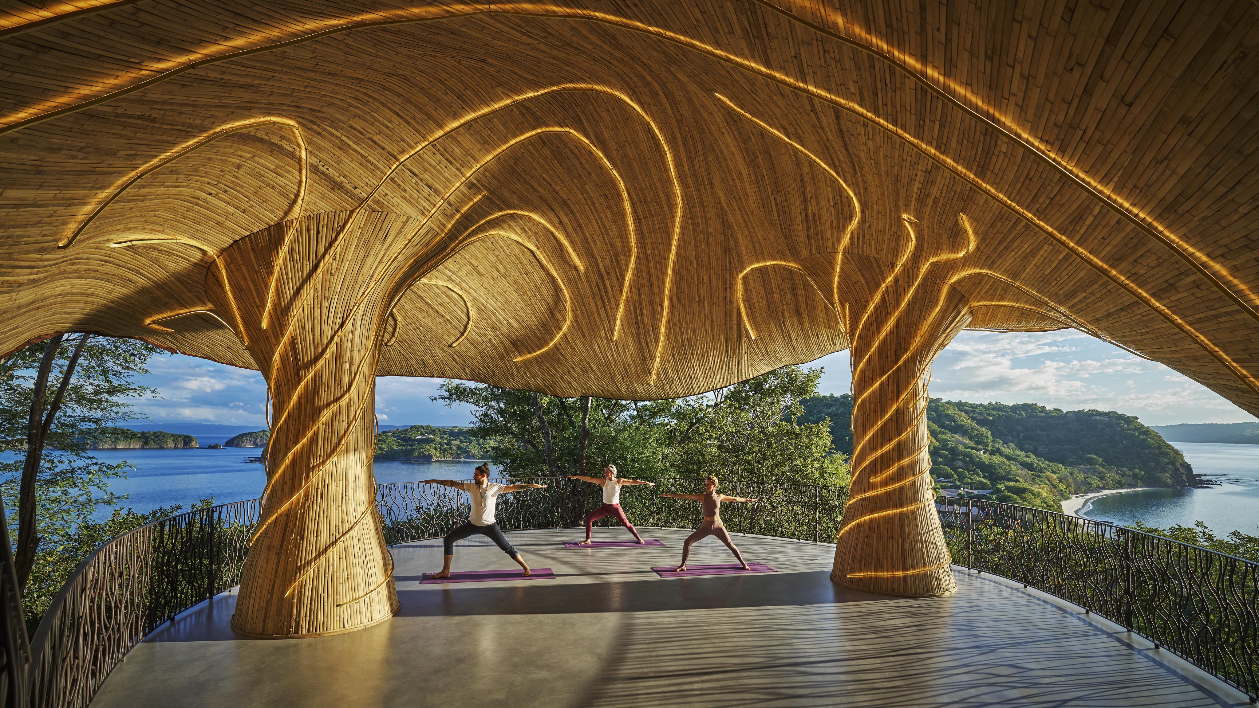 Three people practice yoga under a uniquely designed wooden pavilion in Central America, with a breathtaking view of the ocean and cliffs in the background.