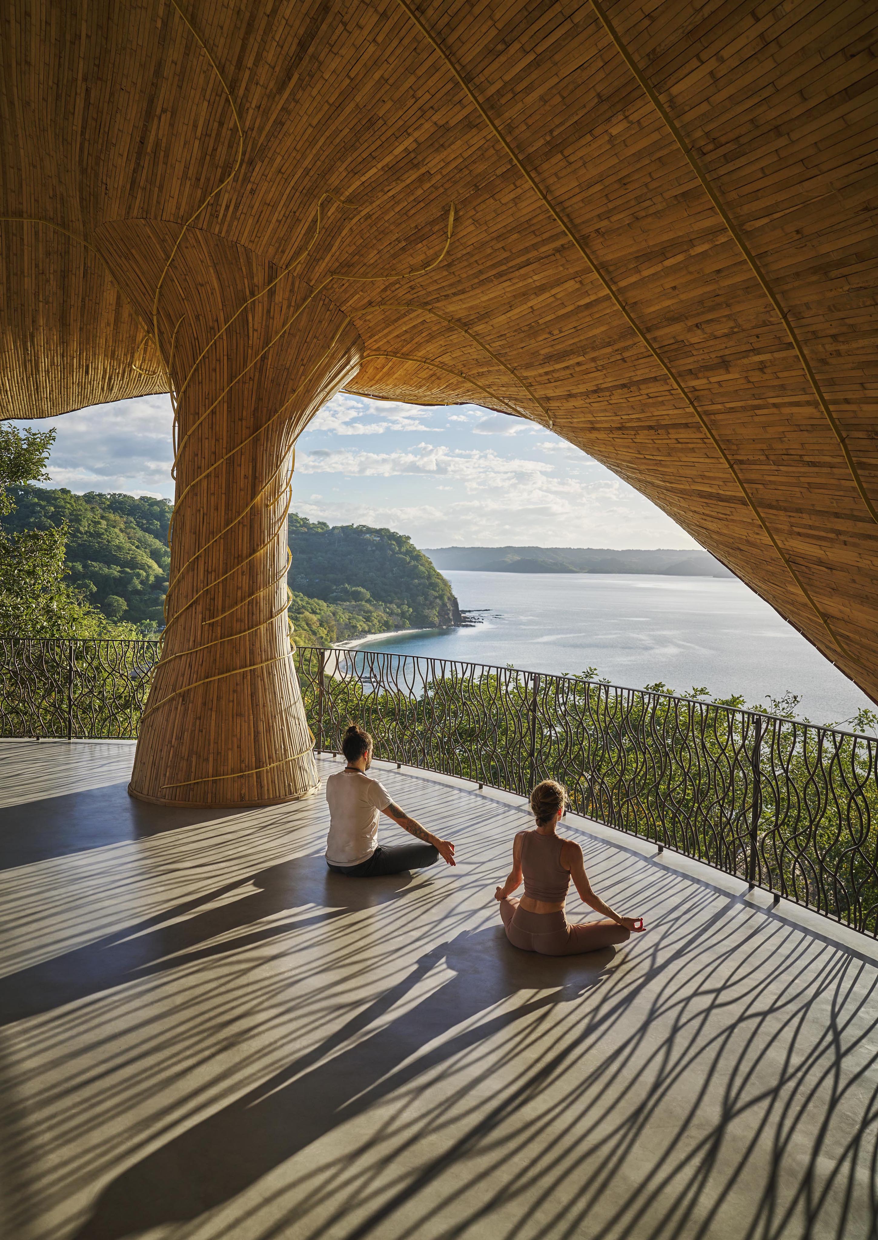 Two people practice yoga on a shaded platform with a large wooden column, overlooking a tranquil body of water and lush greenery under the bright sky, reminiscent of Central America's serene landscapes.