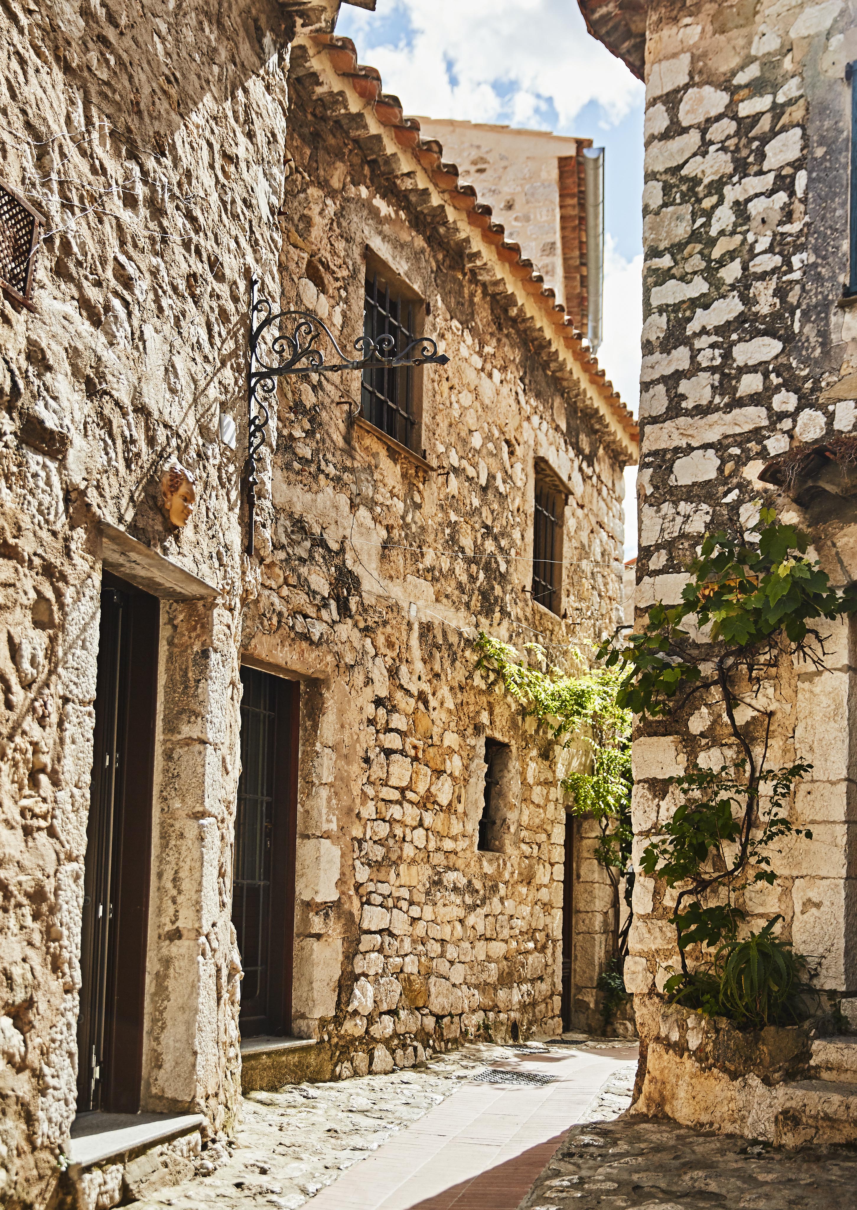 Narrow cobblestone alley lined with weathered stone buildings and greenery climbing the walls, under a partly cloudy blue sky.