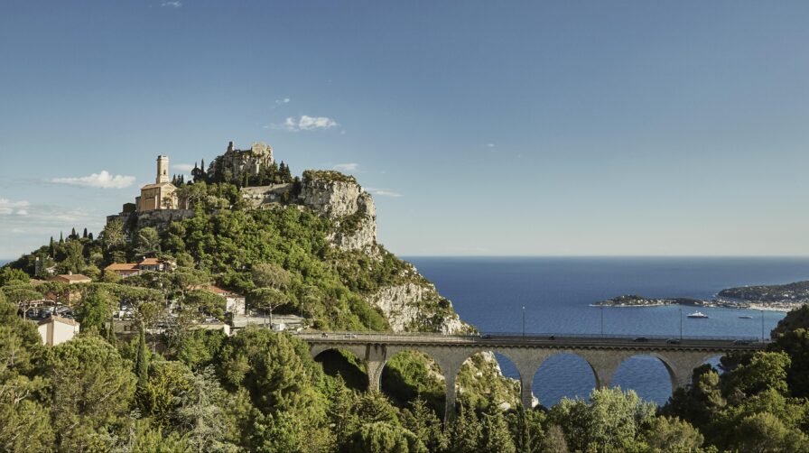 A scenic view of Èze village on a hilltop with a church, surrounded by greenery, overlooking the Mediterranean Sea. A stone bridge arches in the foreground.