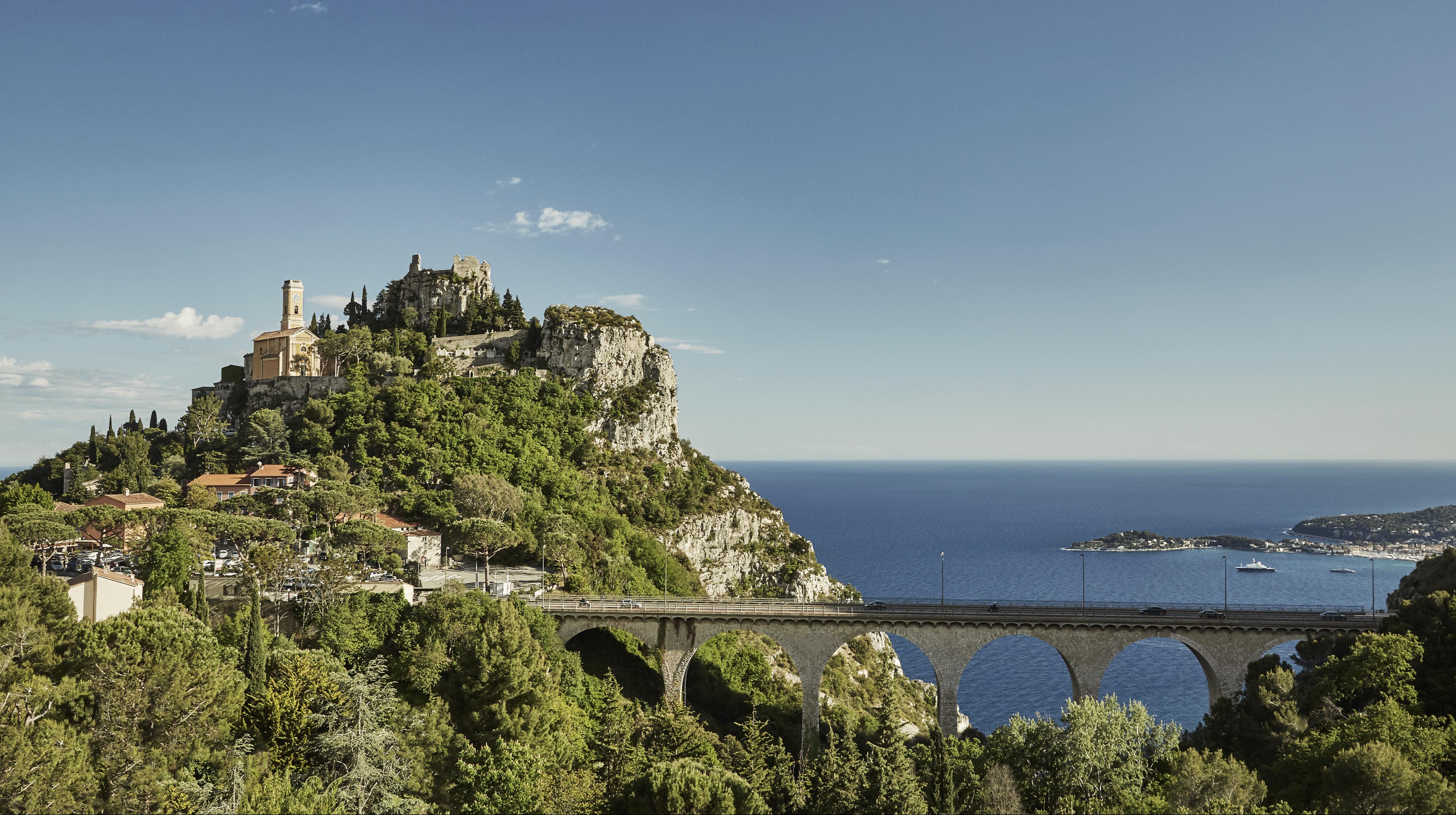 A scenic view of Èze village on a hilltop with a church, surrounded by greenery, overlooking the Mediterranean Sea. A stone bridge arches in the foreground.