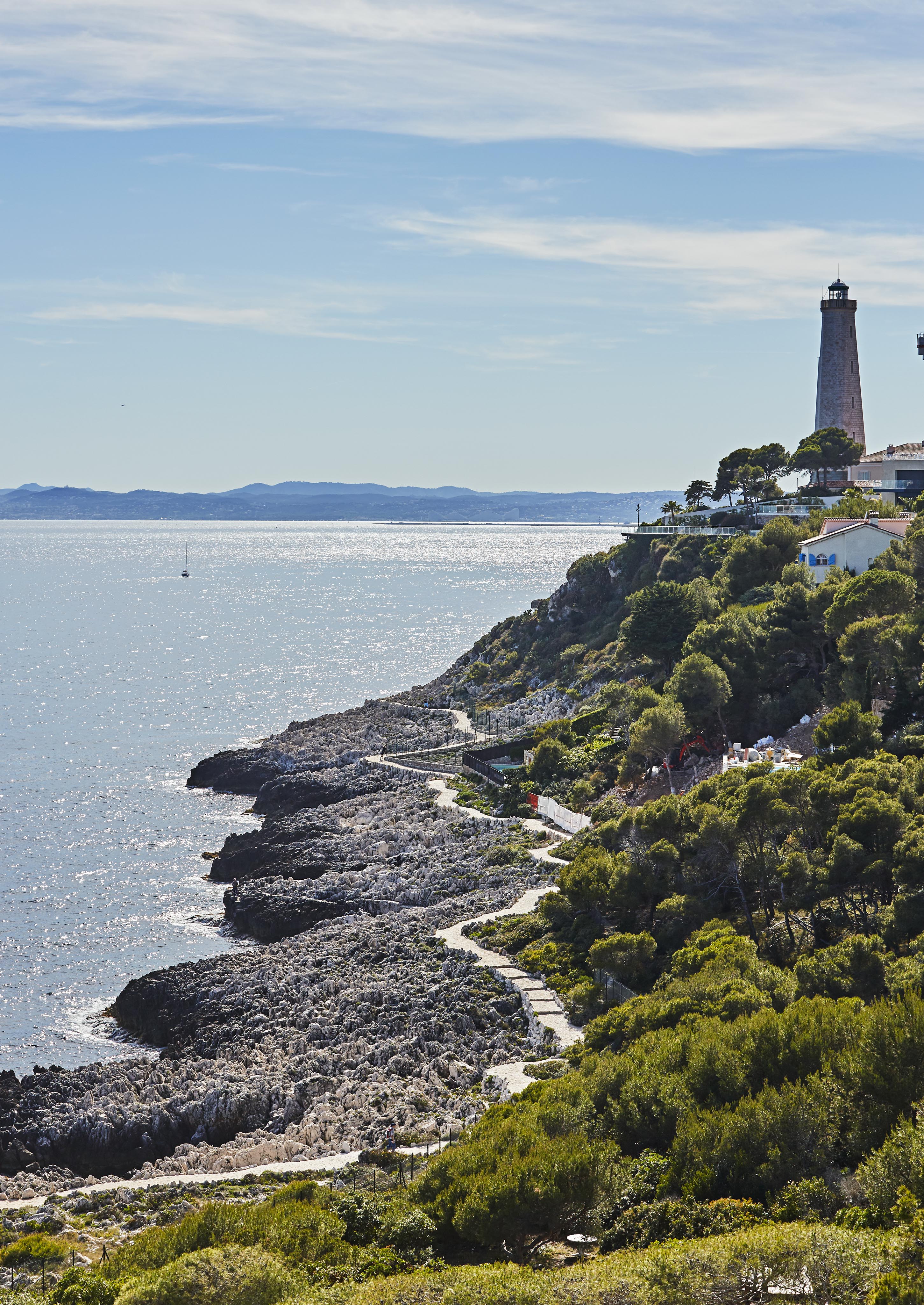 Rocky coastline with a lighthouse on a hilltop, surrounded by trees and overlooking a sparkling sea under a partly cloudy sky.