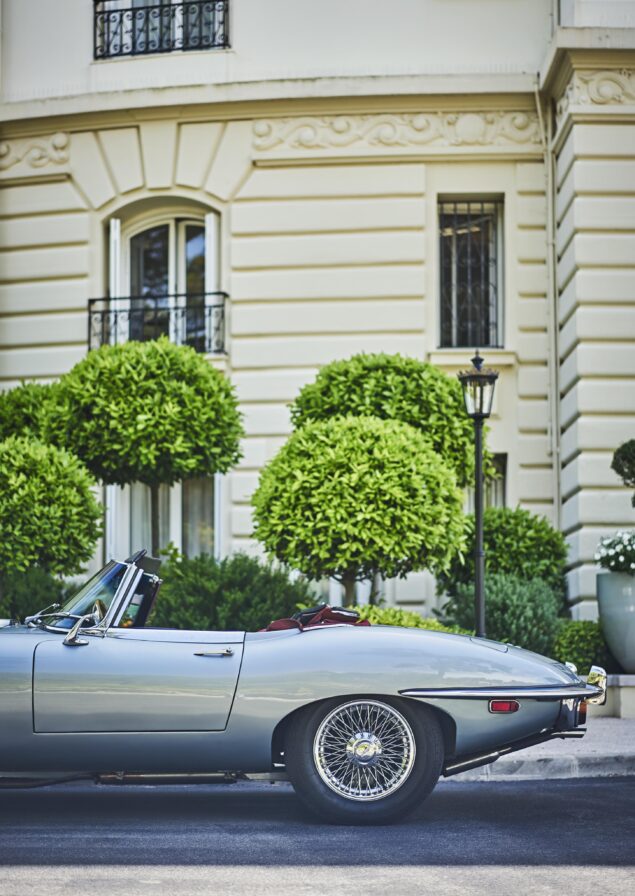 Silver vintage convertible car parked on the street with a beige, ornate building and manicured bushes in the background.