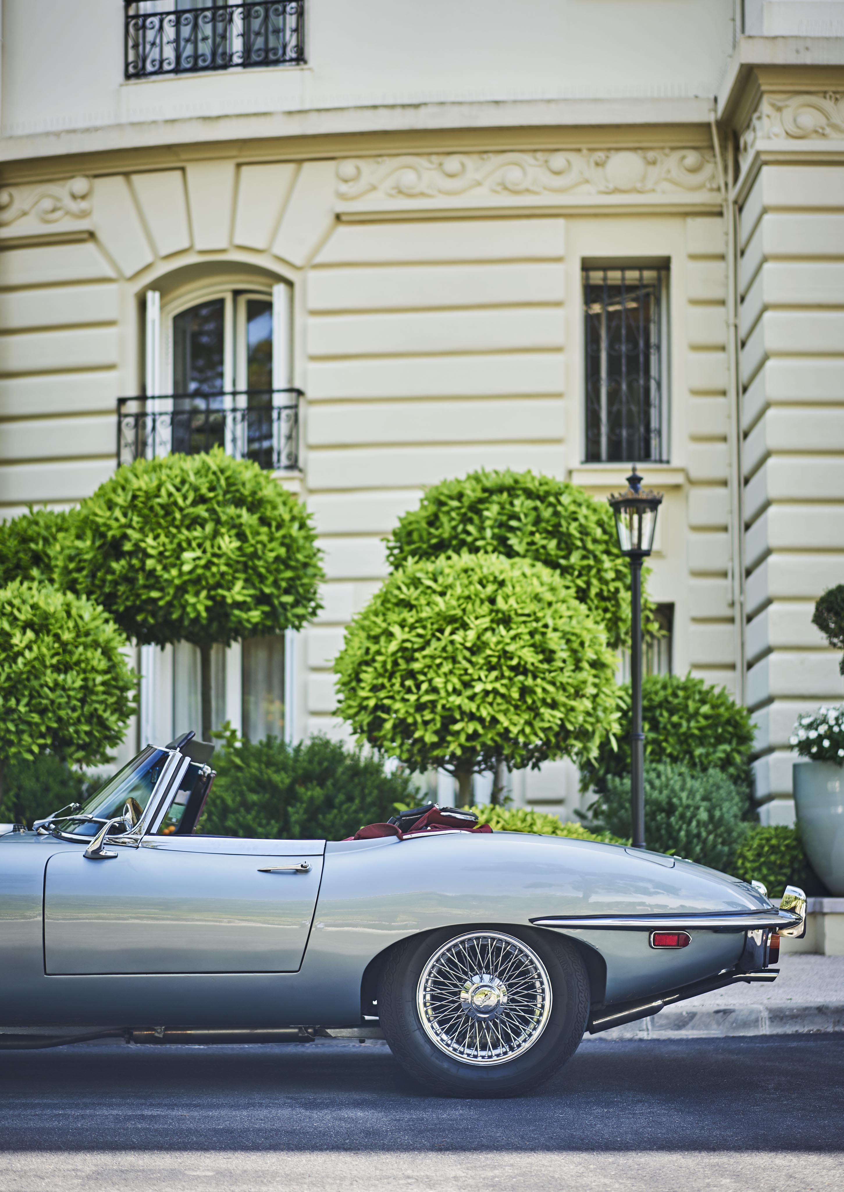 Silver vintage convertible car parked on the street with a beige, ornate building and manicured bushes in the background.