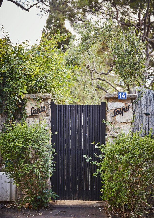 Black vertical slat gate between stone pillars with foliage, marked with the number 14 and words starting with 'Sapia' and 'Soppir'.