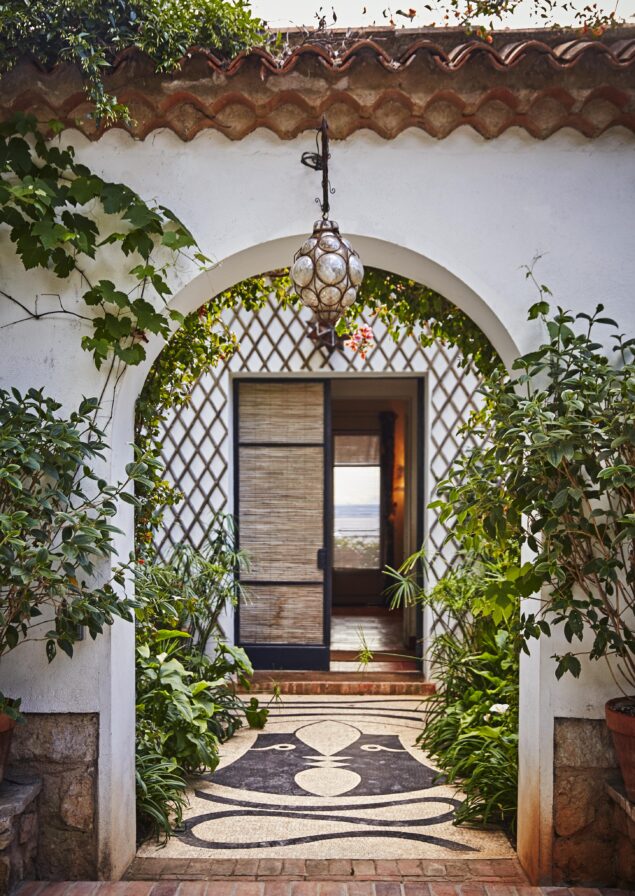 A Mediterranean-style archway leads to a patio with plants, a hanging lantern, and an open door. Tiled floor features a geometric pattern.