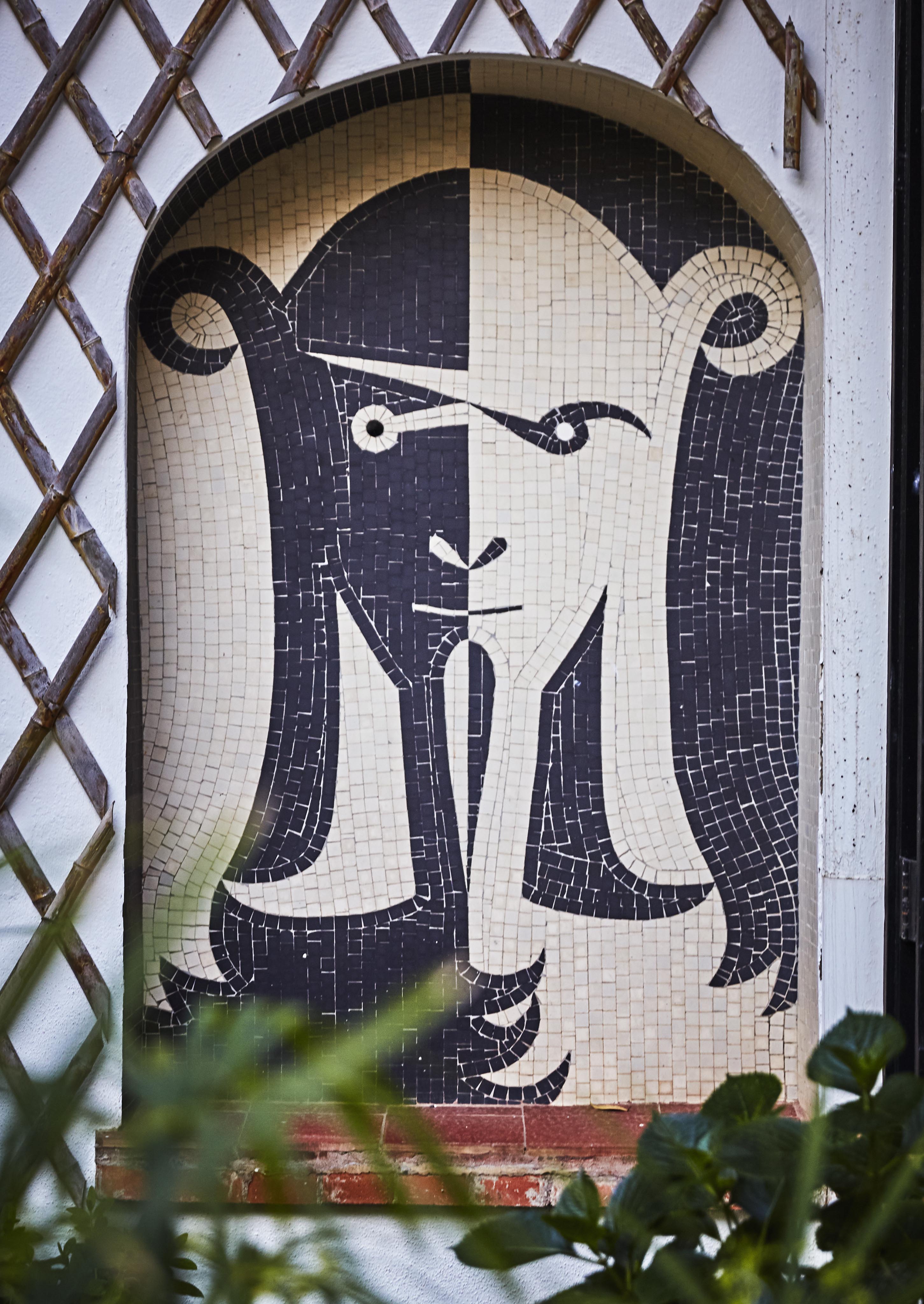 Mosaic art featuring a stylized face in black and white tones, set within an arched niche. Surrounded by a trellis and greenery.